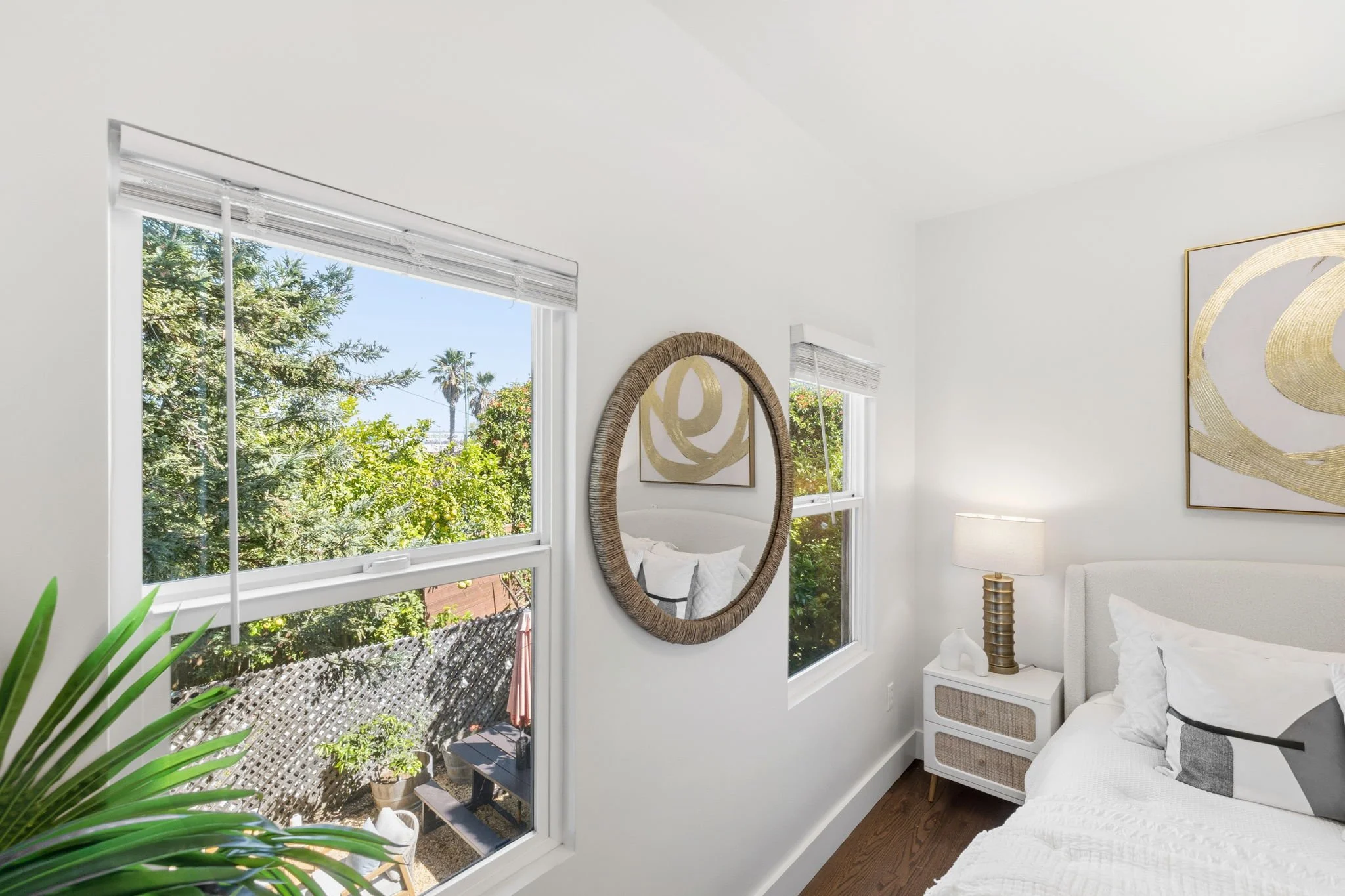 Bright bedroom with two large windows showing green trees and palm trees outside. The room has white walls, a white bed with neutral bedding, a white nightstand with a decorative lamp and colorfully striped vase, and abstract gold and white artwork o