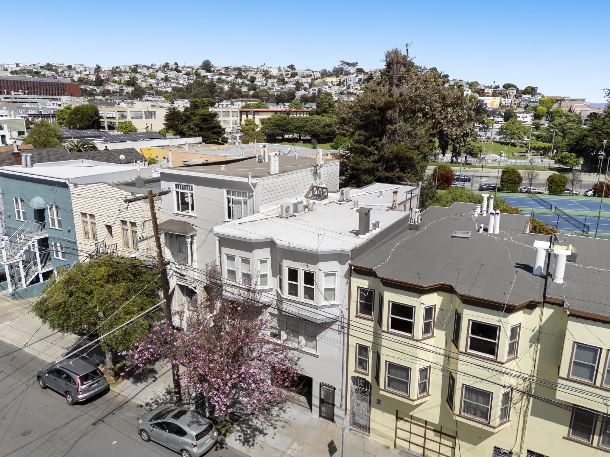 Aerial view of colorful city buildings with rooftops, trees, and a tennis court in the background under a clear blue sky.