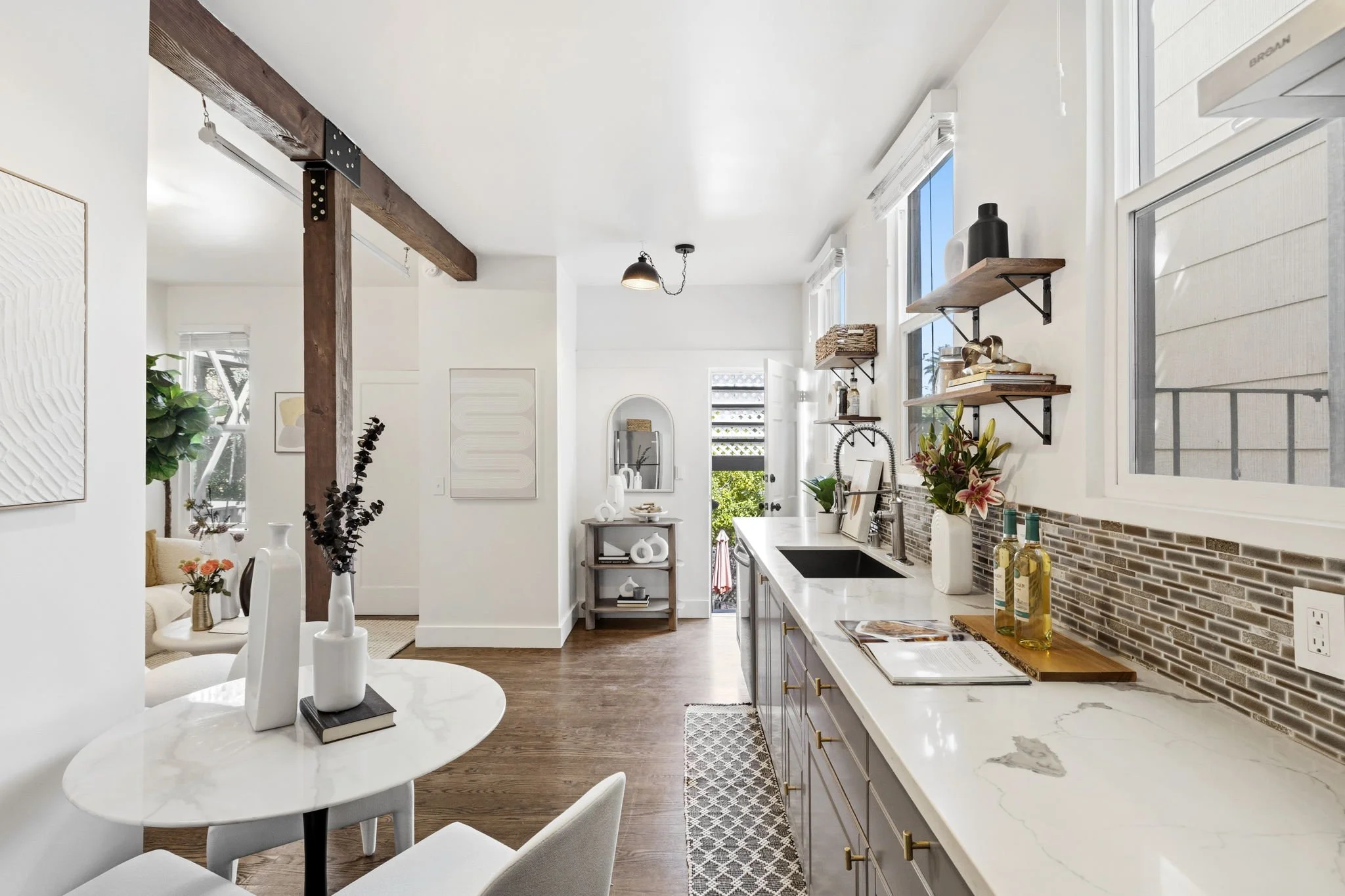 Open-concept kitchen and dining area with white walls, wooden floors, and natural light from windows. Features a marble countertop, open wooden shelves with decor, and a small table with vases and books.
