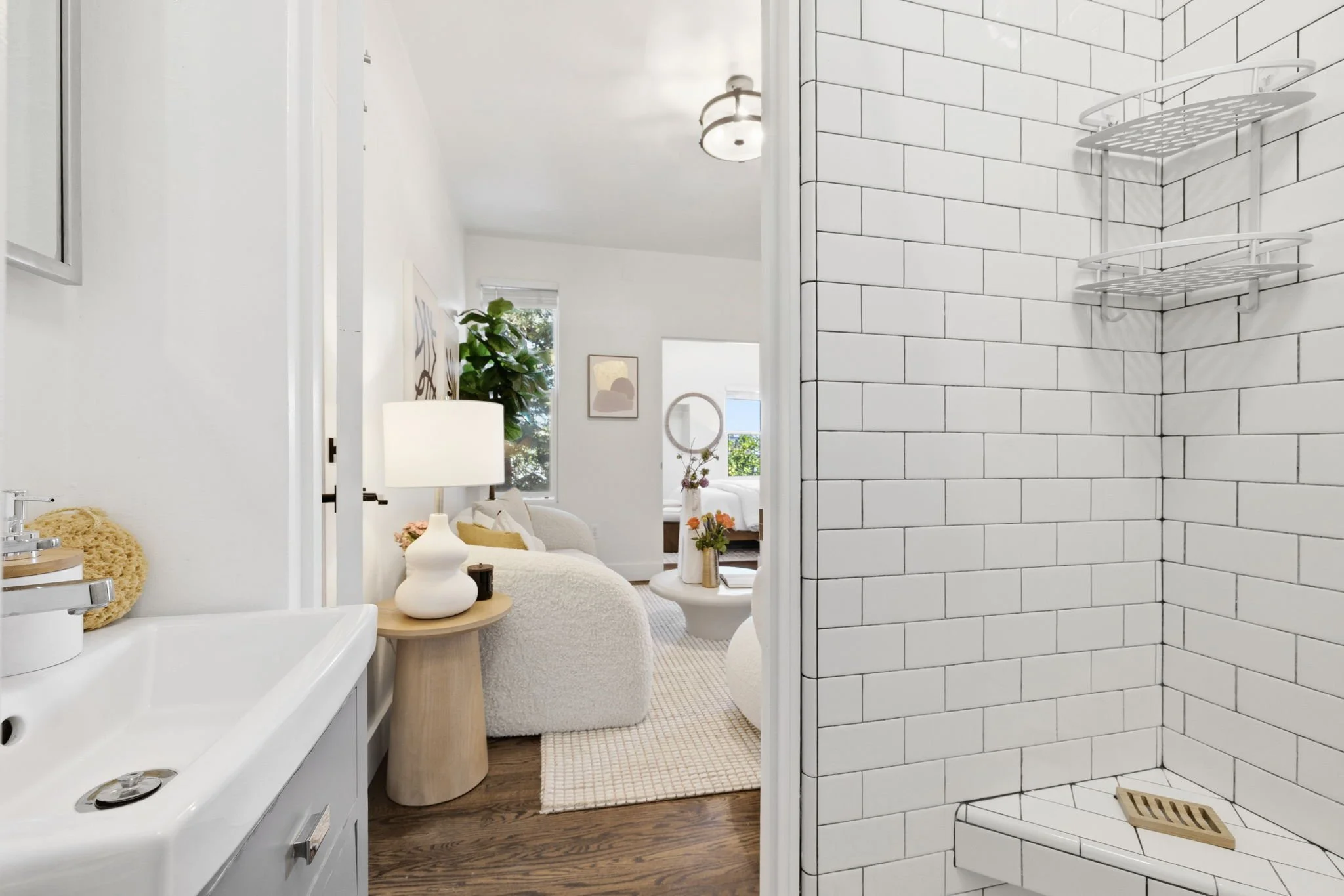 View from a bathroom shower into a living room with a white sofa, a side table, a lamp, plants, and wall art in a bright, modern interior.