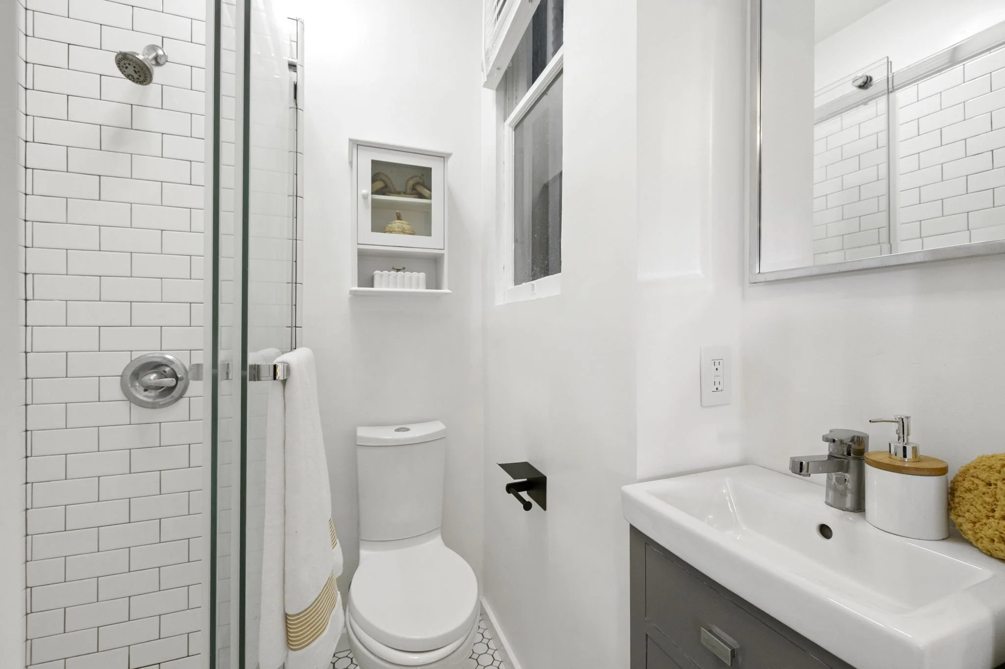 Small modern bathroom with white subway tile in the shower, a white toilet, a small gray vanity with a white sink, a large mirror, a window, and decorative items like a sponge, soap dispenser, and a small shelf with toiletries.