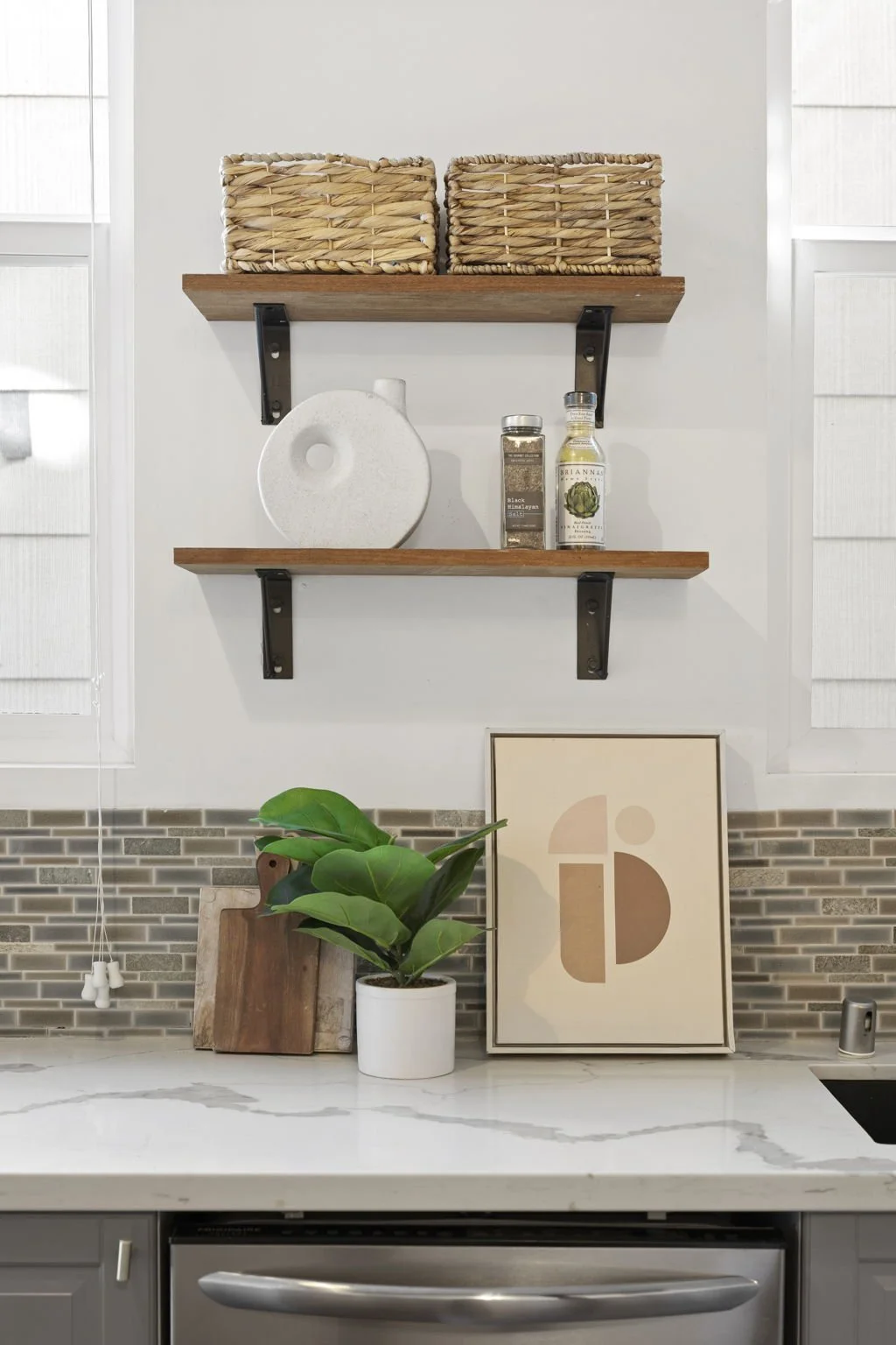 Interior kitchen shelf with two wicker baskets on top, a white ceramic circular object, and two spice jars on a white wall with a ledge, next to a green potted plant, framed abstract art, and a gray countertop.