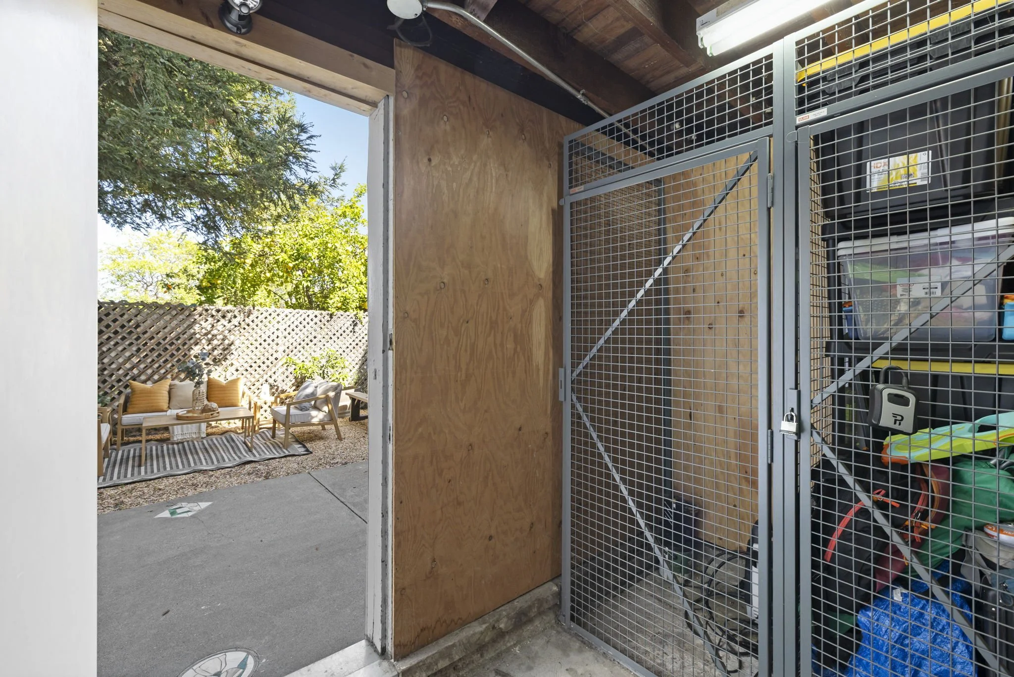 View of a backyard patio with outdoor furniture and a lattice fence, seen through an open door from a storage shed that contains organized tools and supplies.