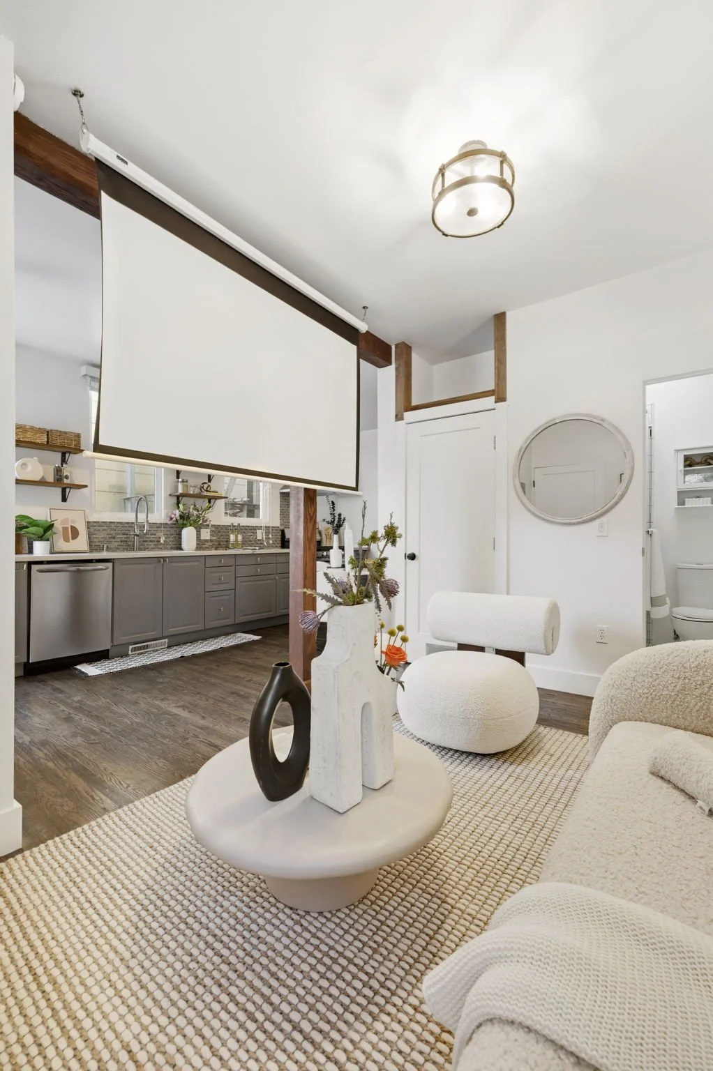 Living room with a white sofa, round ottoman, decorative vases on a coffee table, and a large pull-down projection screen, overlooking the kitchen with gray cabinets and stainless steel appliances.