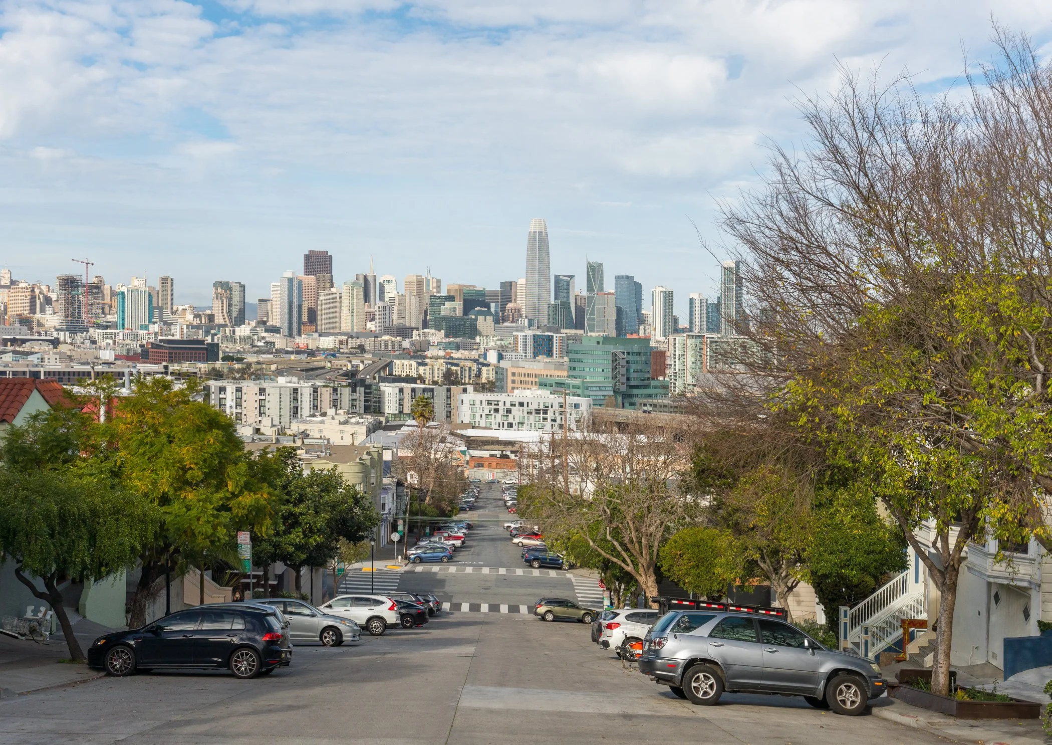 View of a city skyline with a mix of modern skyscrapers and shorter buildings, seen from a residential street with parked cars and trees in the foreground.