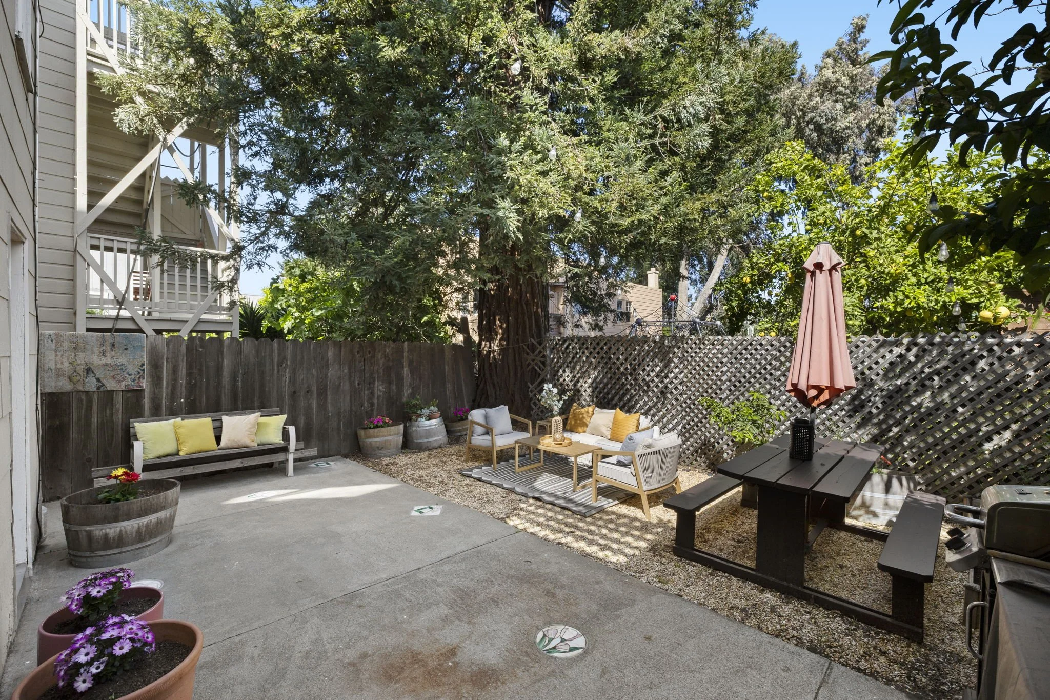 A cozy backyard patio with various seating options, potted flowers, and a large tree providing shade, enclosed by wooden fences.
