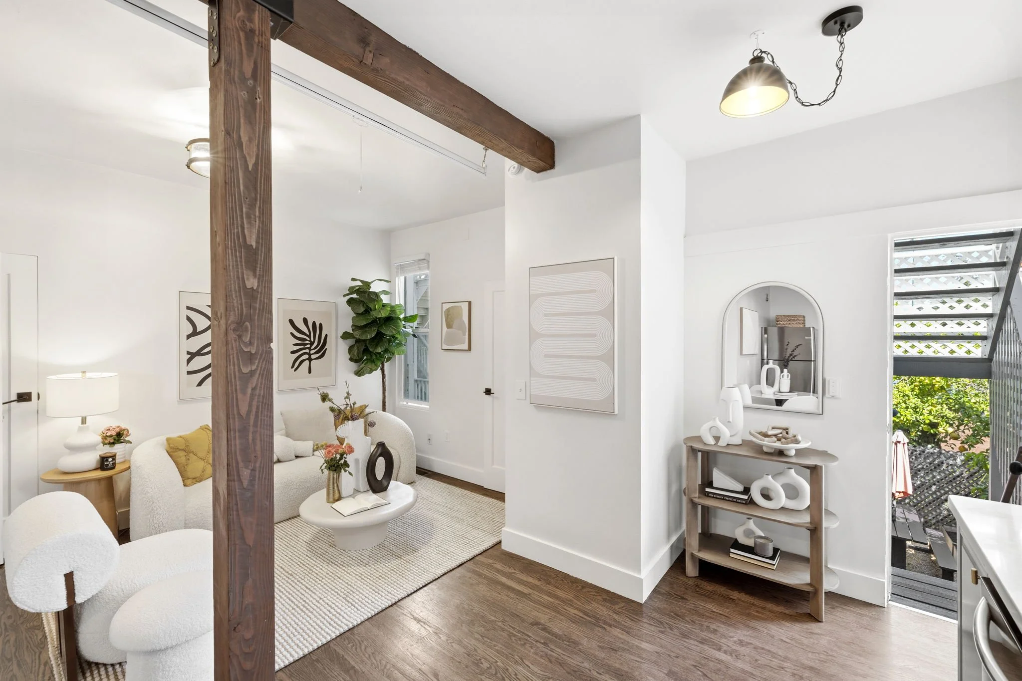 A bright, modern living room with white walls, wooden flooring, and minimalist decor. A white couch with yellow cushion, a small coffee table with vases, and abstract artwork on the walls. There's a tall green plant near a window, a side table with a