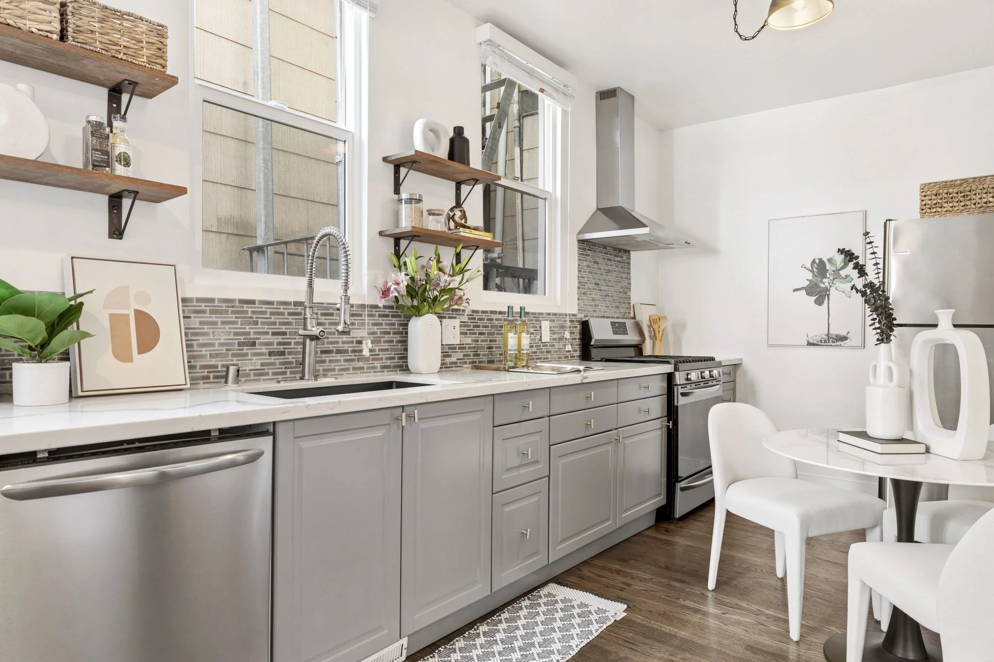 Modern kitchen with gray cabinets, stainless steel appliances, open wooden shelves, a white marble countertop, and decorative vases and plants. There is a small dining area with white chairs and a round table.