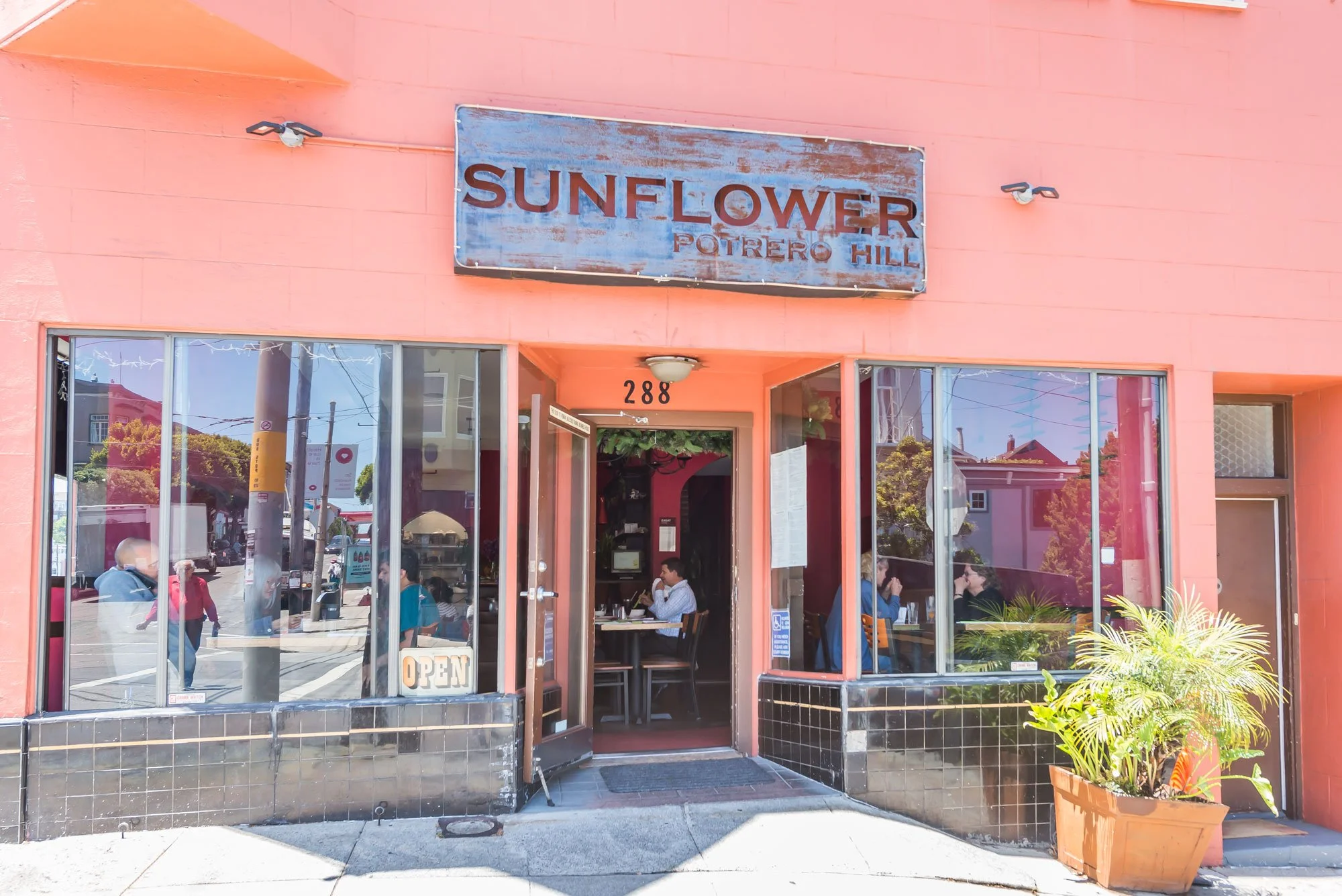 The exterior of a restaurant called Sunflower Potrero Hill, with pink walls, large glass windows, and an open door. There are customers inside, and a potted plant outside near the entrance.
