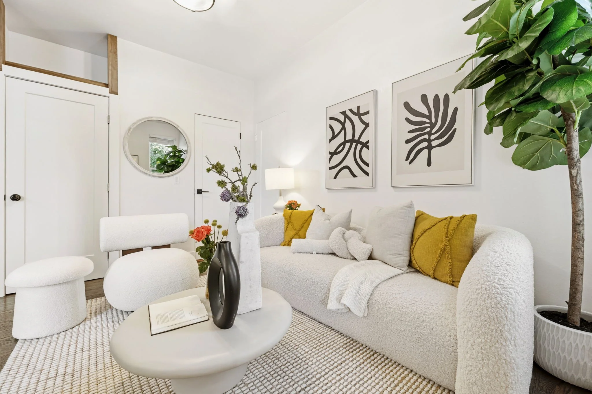 Living room with white textured sofa, yellow and white pillows, abstract black and white framed artwork, large potted plant, round white coffee table with vases and open book, white walls, door with round mirror, lamp, and decorative branch in vase.