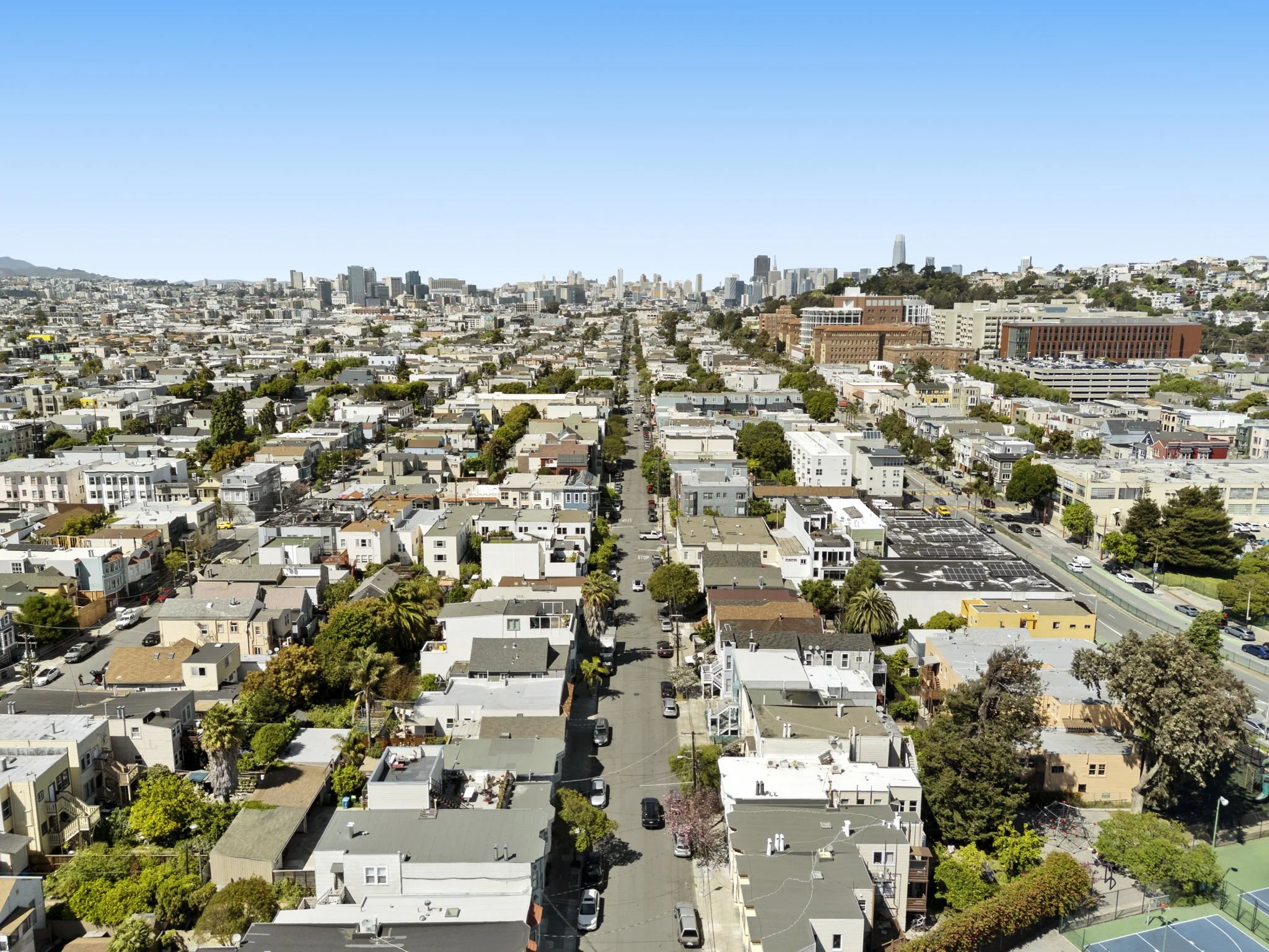 Aerial view of a city neighborhood with residential houses, trees, and streets, with a downtown skyline in the background under a clear blue sky.