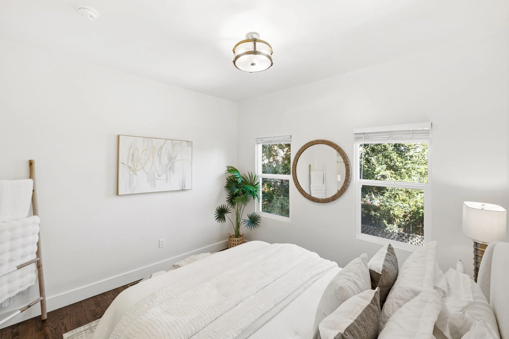 Bright bedroom with white walls, a bed with white bedding and pillows, two windows with views of green trees, a round mirror on the wall, a potted plant, a modern ceiling light, and a lamp on a nightstand.