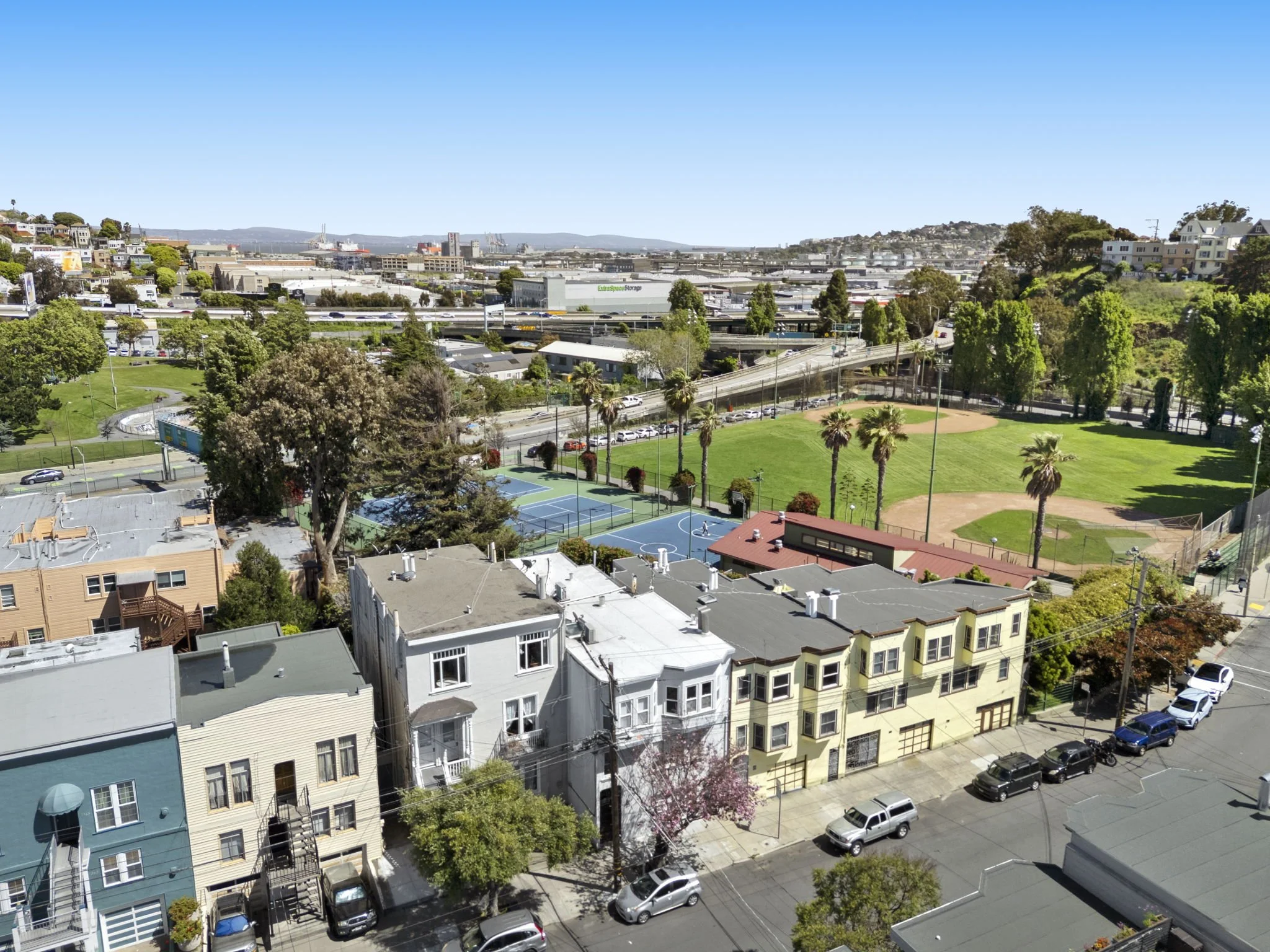 Aerial cityscape showing houses, a park with tennis courts and baseball field, and busy highway with traffic under a clear blue sky.