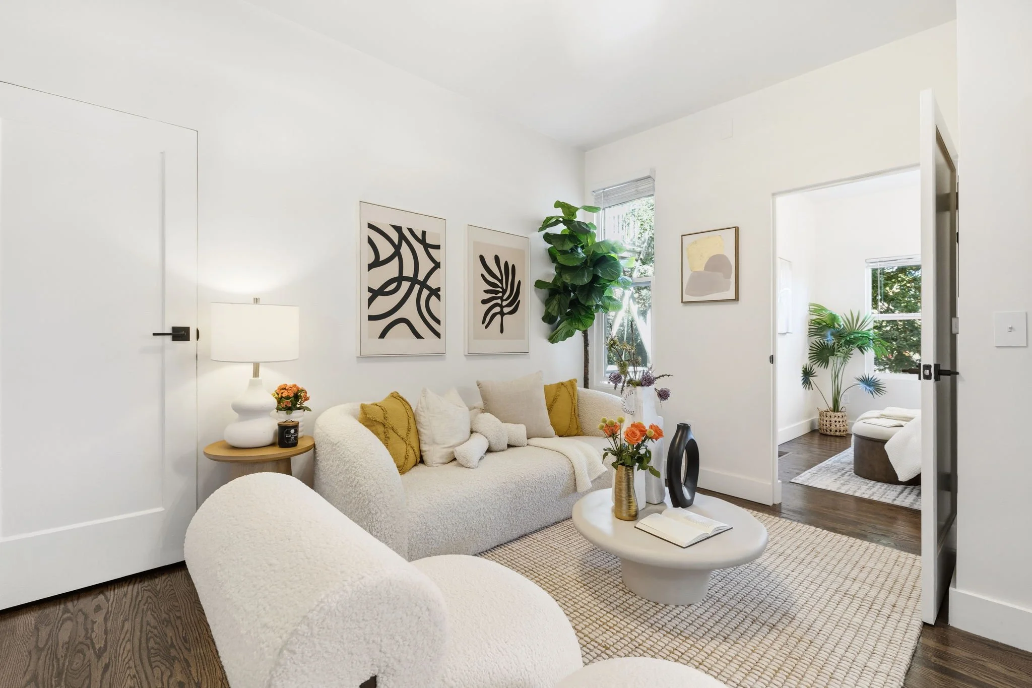 Bright living room with white walls, beige sofa with mustard and white cushions, black and white artwork, large plant by the window, white armchair, round white coffee table with vases and books, wooden side table with lamp and flowers, doorway to another room with a potted plant.