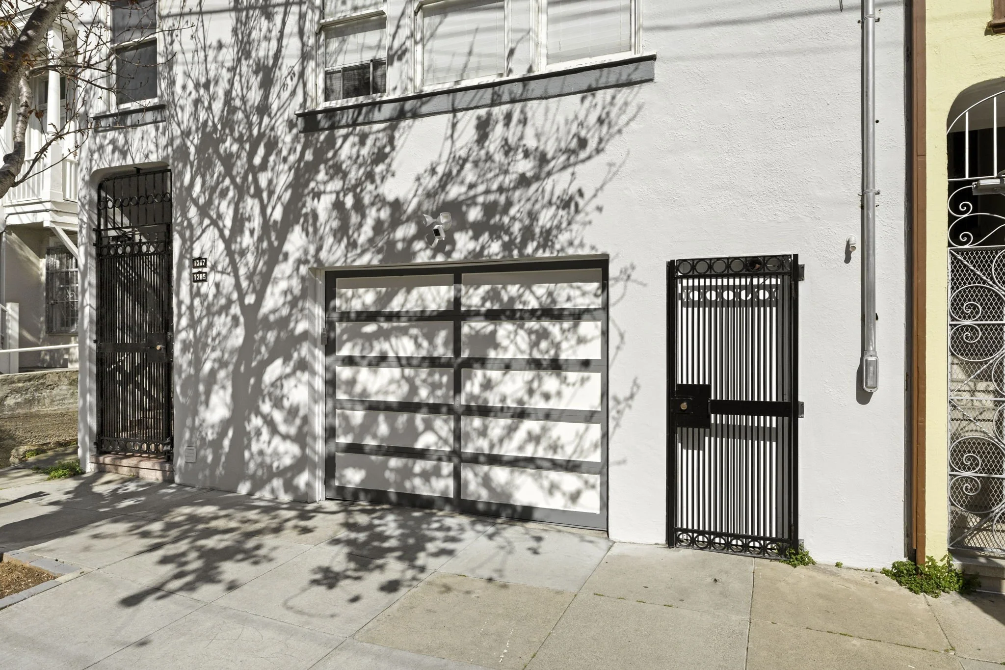 White building exterior with two black metal gates and a parked gray garage door. Shadow of a tree cast on the wall.