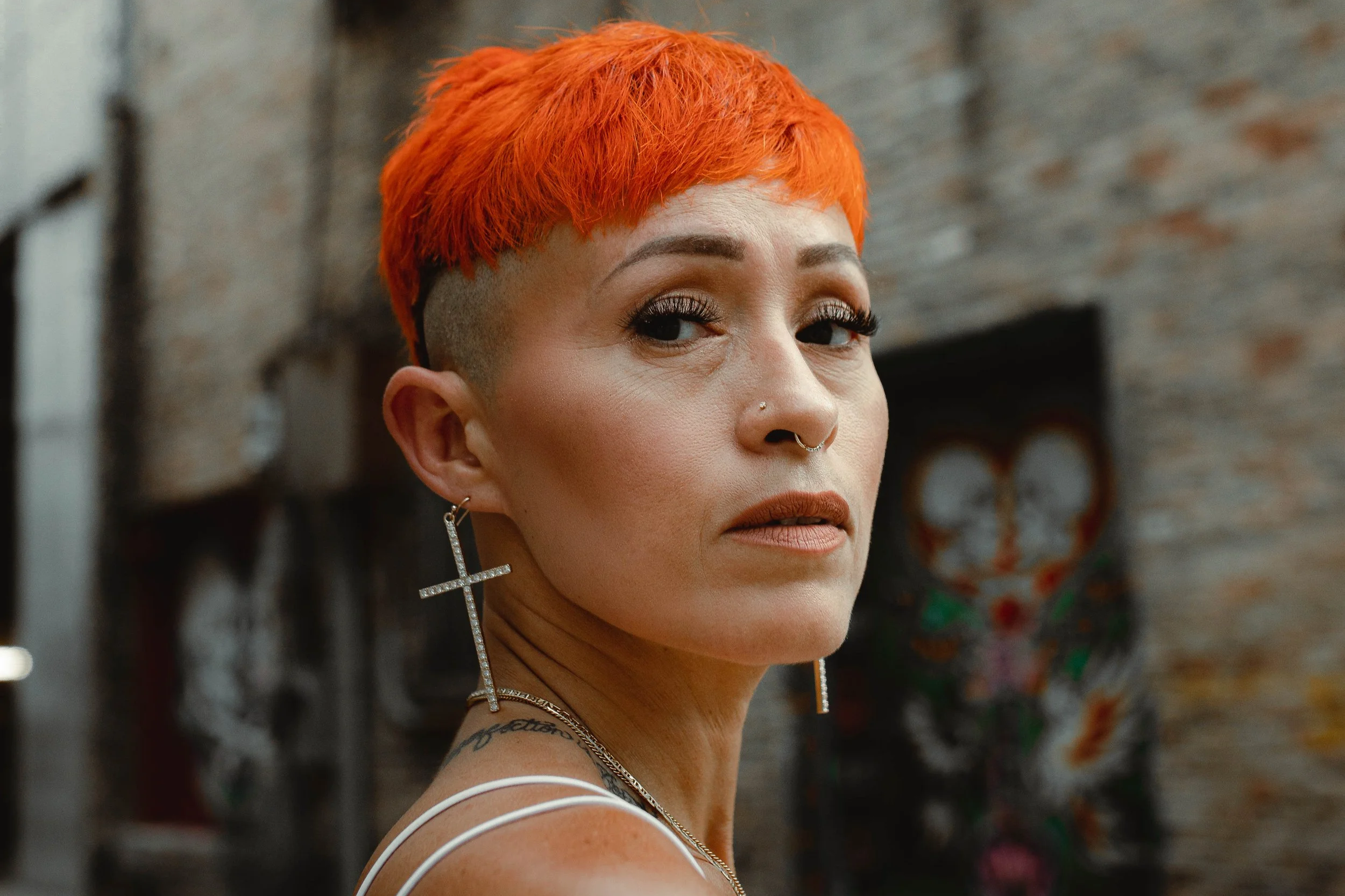 A woman with short bright orange hair, wearing large cross earrings and makeup, standing in front of graffiti on a brick wall.