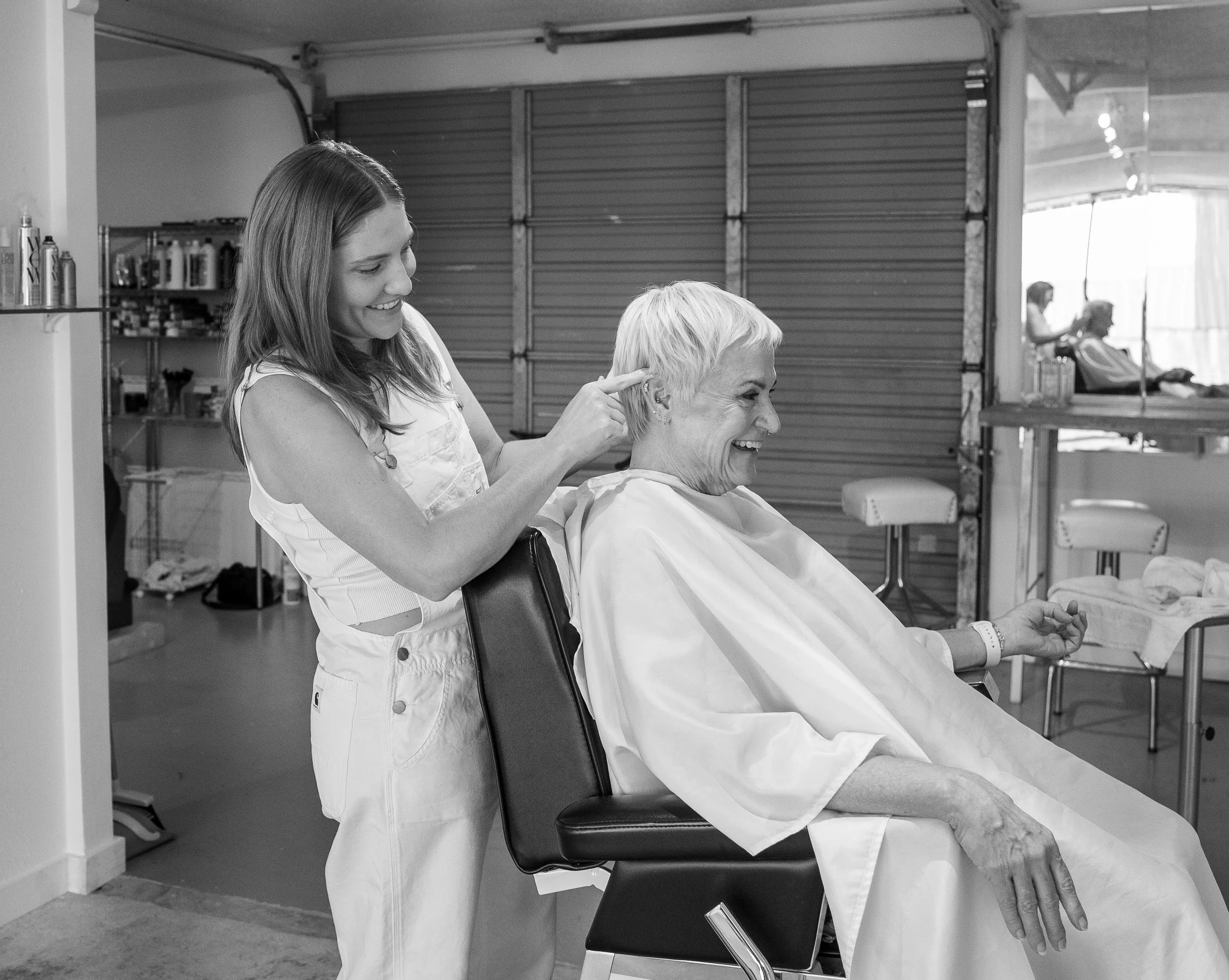 A young woman shampooing an older woman's hair at a hair salon, both smiling.
