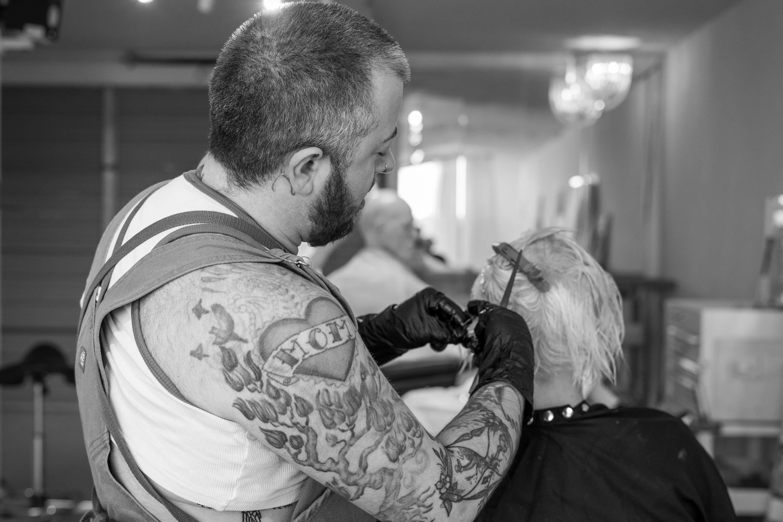 A tattooed man with a beard and a nose ring is getting a haircut in a salon, while seated in a barber chair. There are chandeliers hanging from the ceiling in the background.