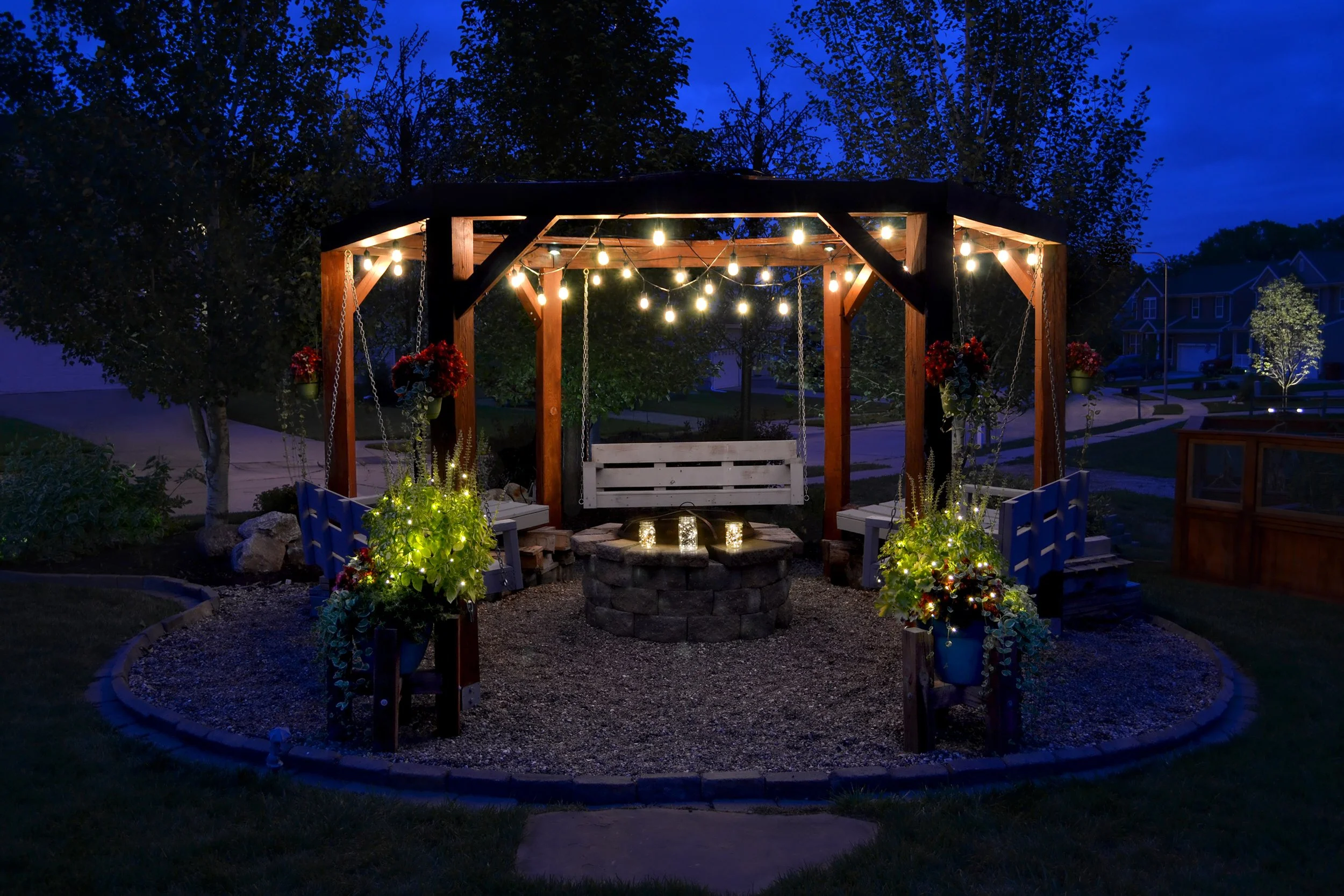 Nighttime outdoor scene featuring a wooden porch swing under a pergola decorated with string lights. Potted plants with flowers are around the base, and the background includes trees and houses illuminated by outdoor lighting.