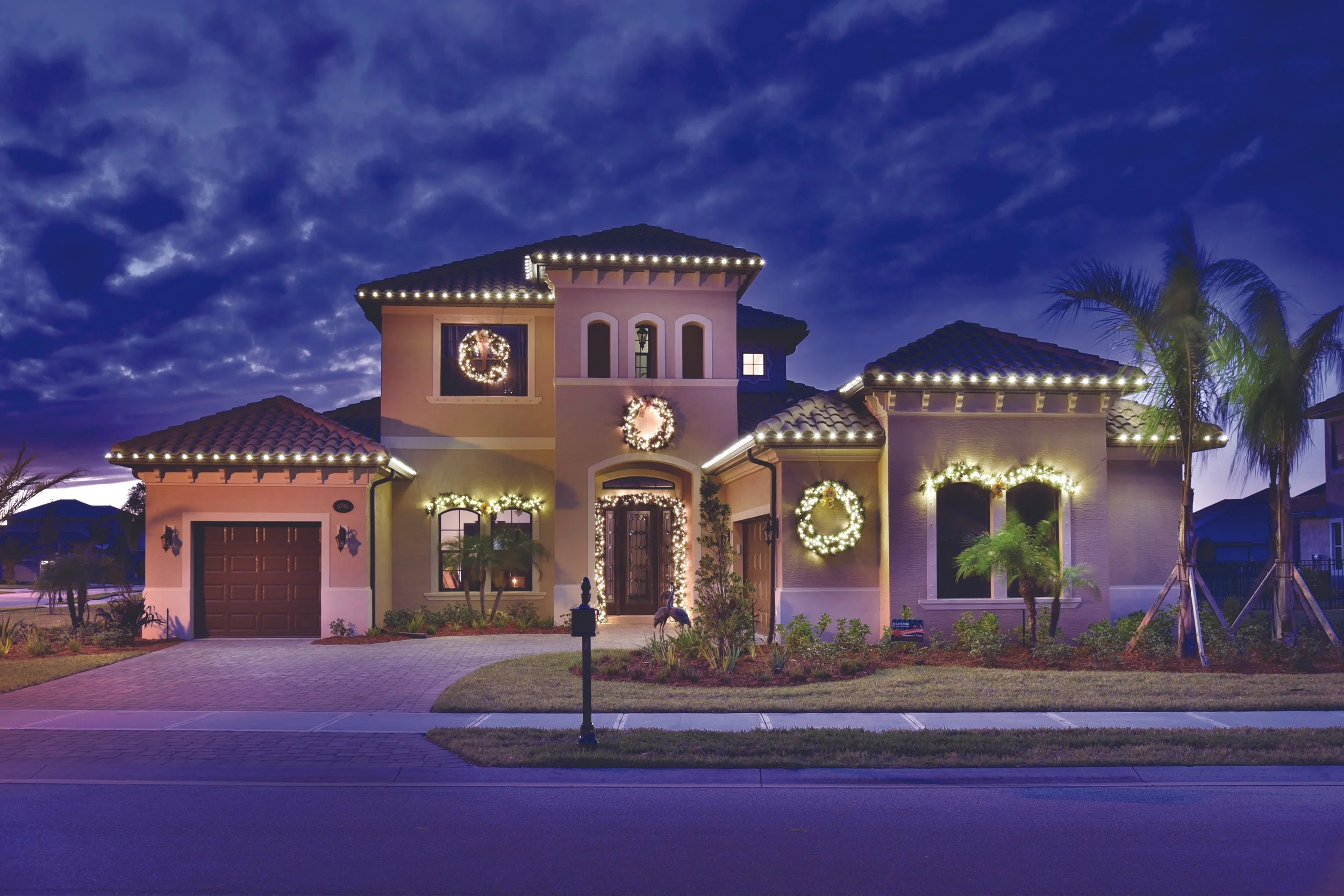 Night view of a house decorated with Christmas lights and wreaths, with the sky filled with clouds and a palm tree on the right side.