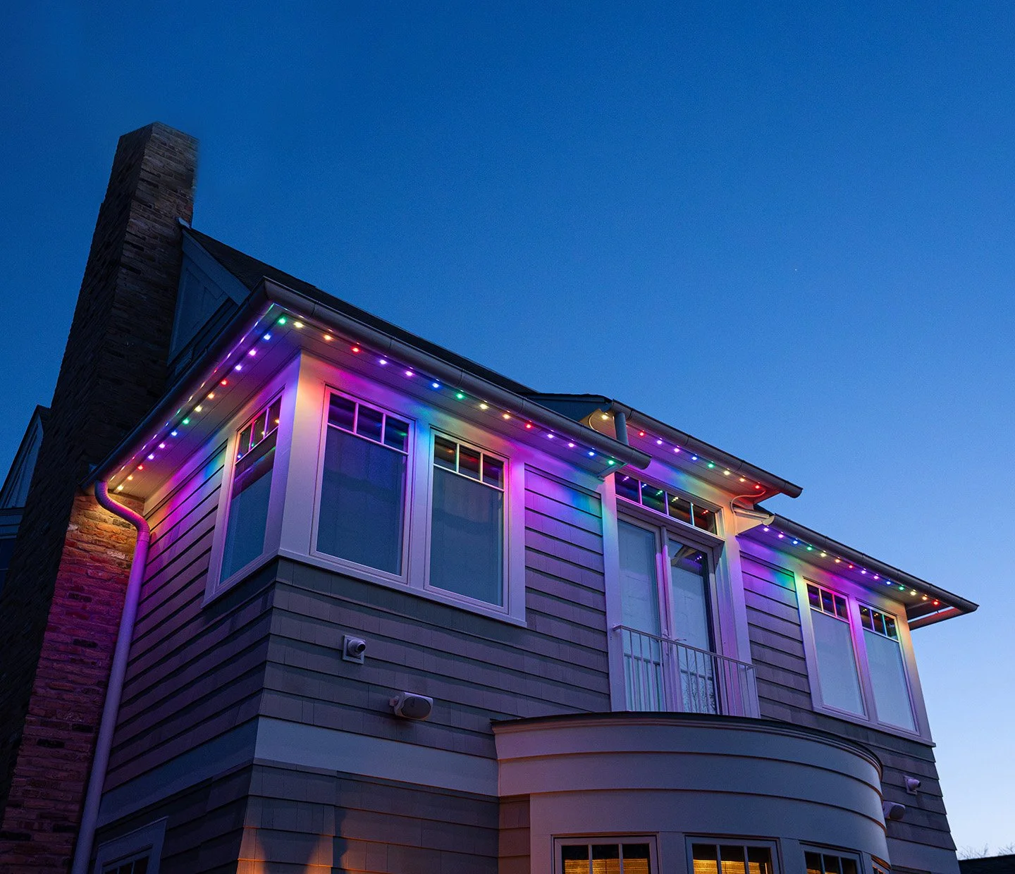 A house illuminated with colorful string lights at dusk, showcasing an upper floor with large windows and a chimney on the side.