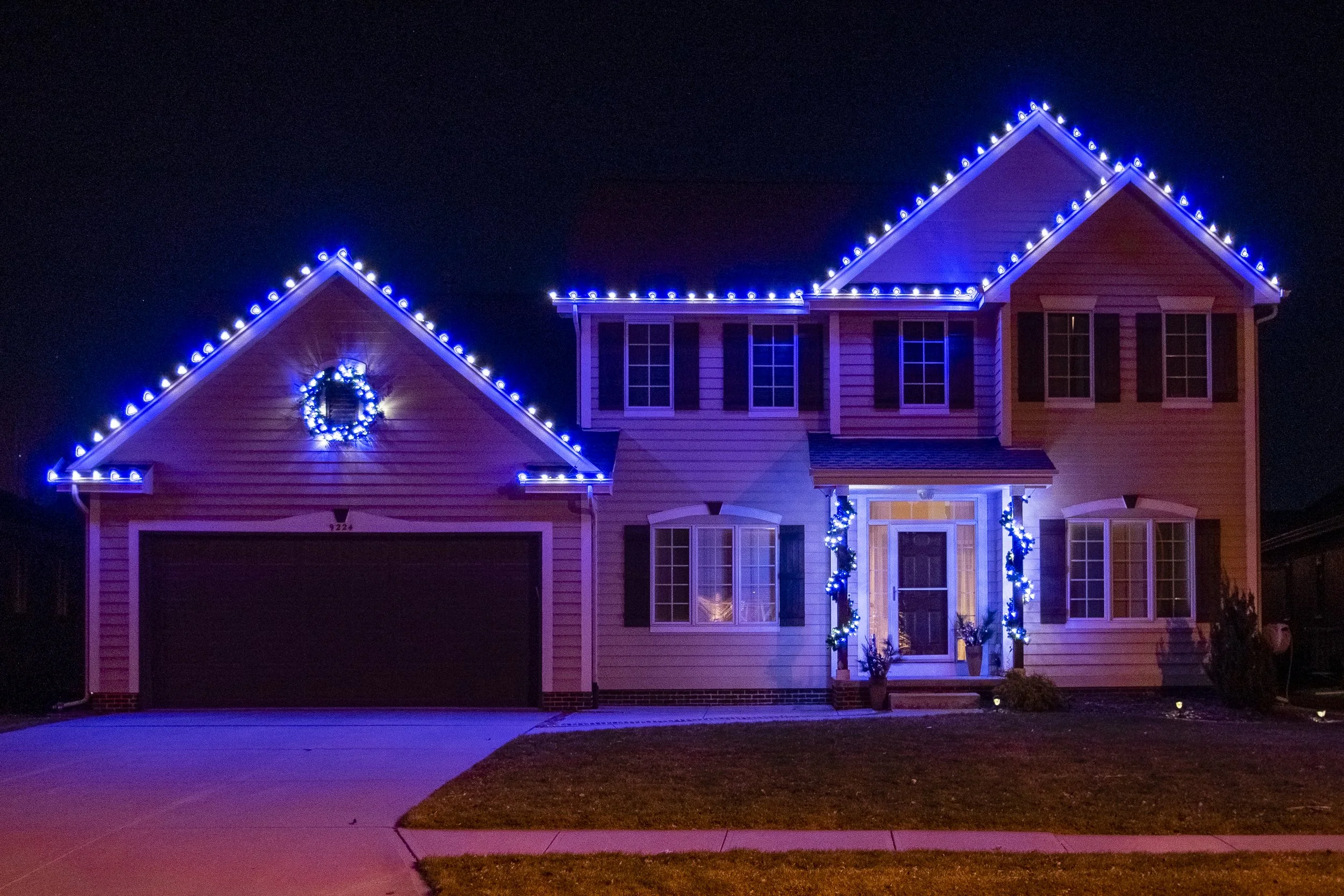 A two-story house decorated with blue and white Christmas lights outlining the roof and windows at night. There is a wreath on the garage door and garland around the front door.