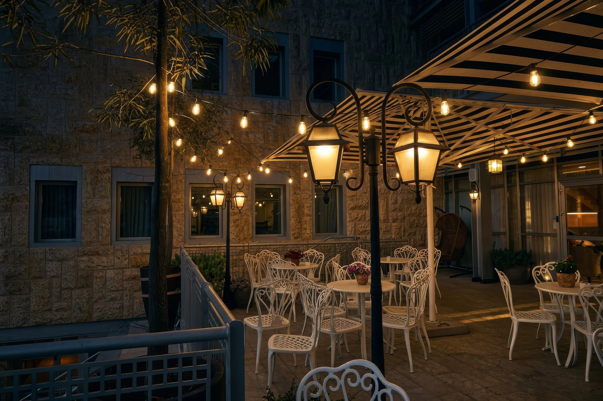 Outdoor patio or terrace area with white metal chairs, lit string lights, and lantern-style lamps hanging from poles, with a building with stone walls and several windows in the background.