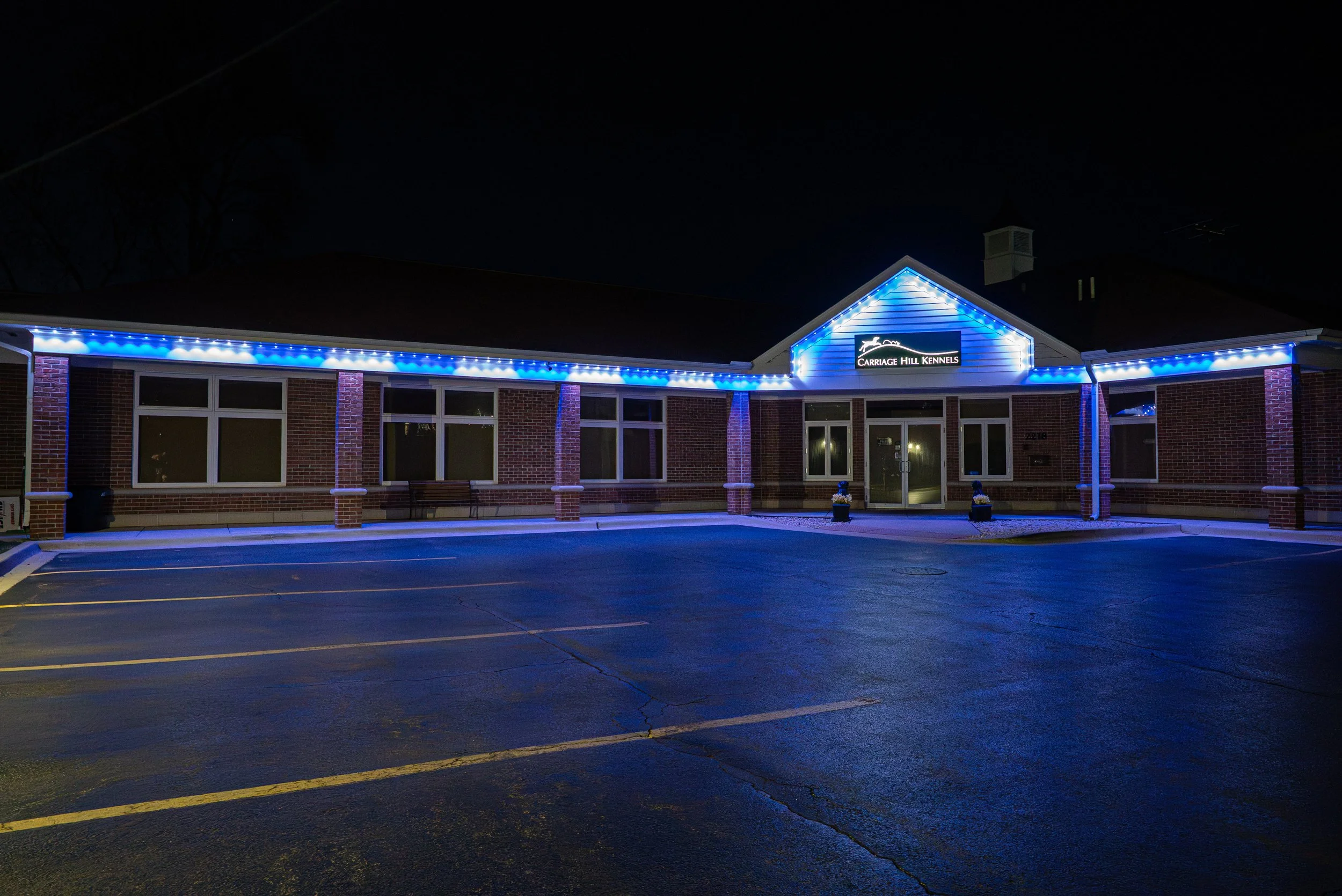 Night view of a building with a sign that reads 'Carriage Hill Kennels,' illuminated with blue lights along the roofline and around the sign. The building has brick walls, multiple windows, and a parking lot in the foreground.