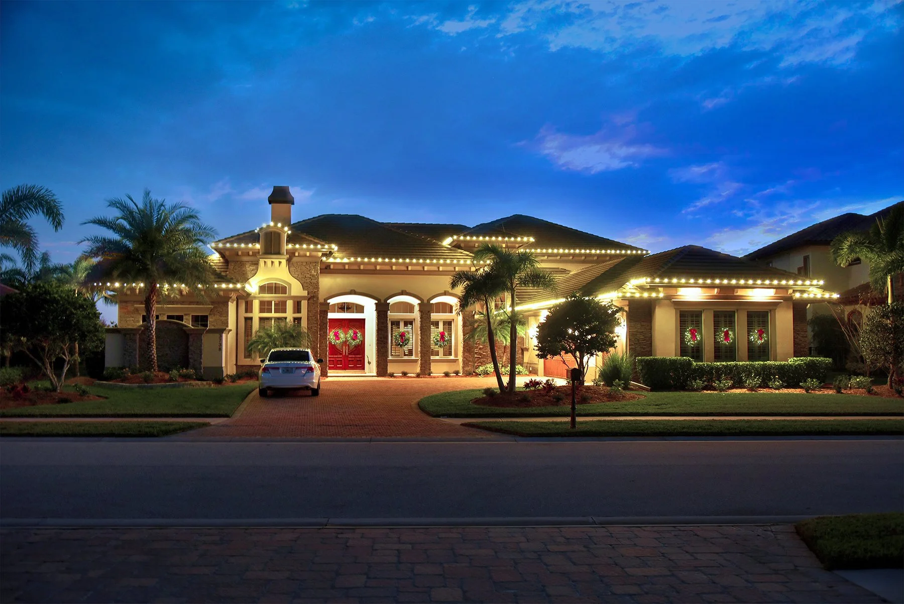 A large house decorated with Christmas wreaths on the windows and a wreath on the front door, with white string lights outlining the roof. The house is surrounded by palm trees, bushes, and a small front yard. The driveway has a parked car, and the sky is dusk with some clouds visible.