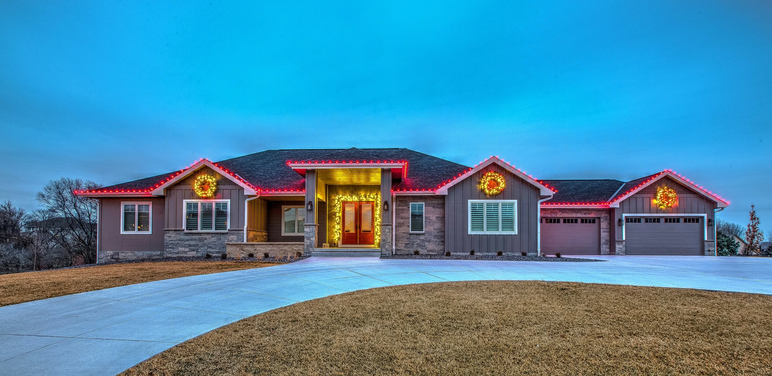 Nighttime view of a house with brick walls and white-framed arched windows decorated with Christmas wreaths. The windows reveal a warmly lit interior with Christmas decorations and a Christmas tree. There is snow and plants below the windows.