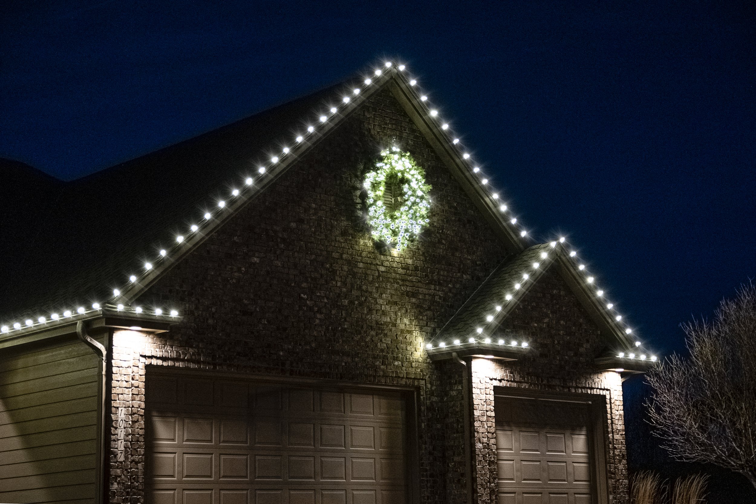 A brick house decorated with white Christmas lights on the roof and a green wreath with white accents on the front of the house