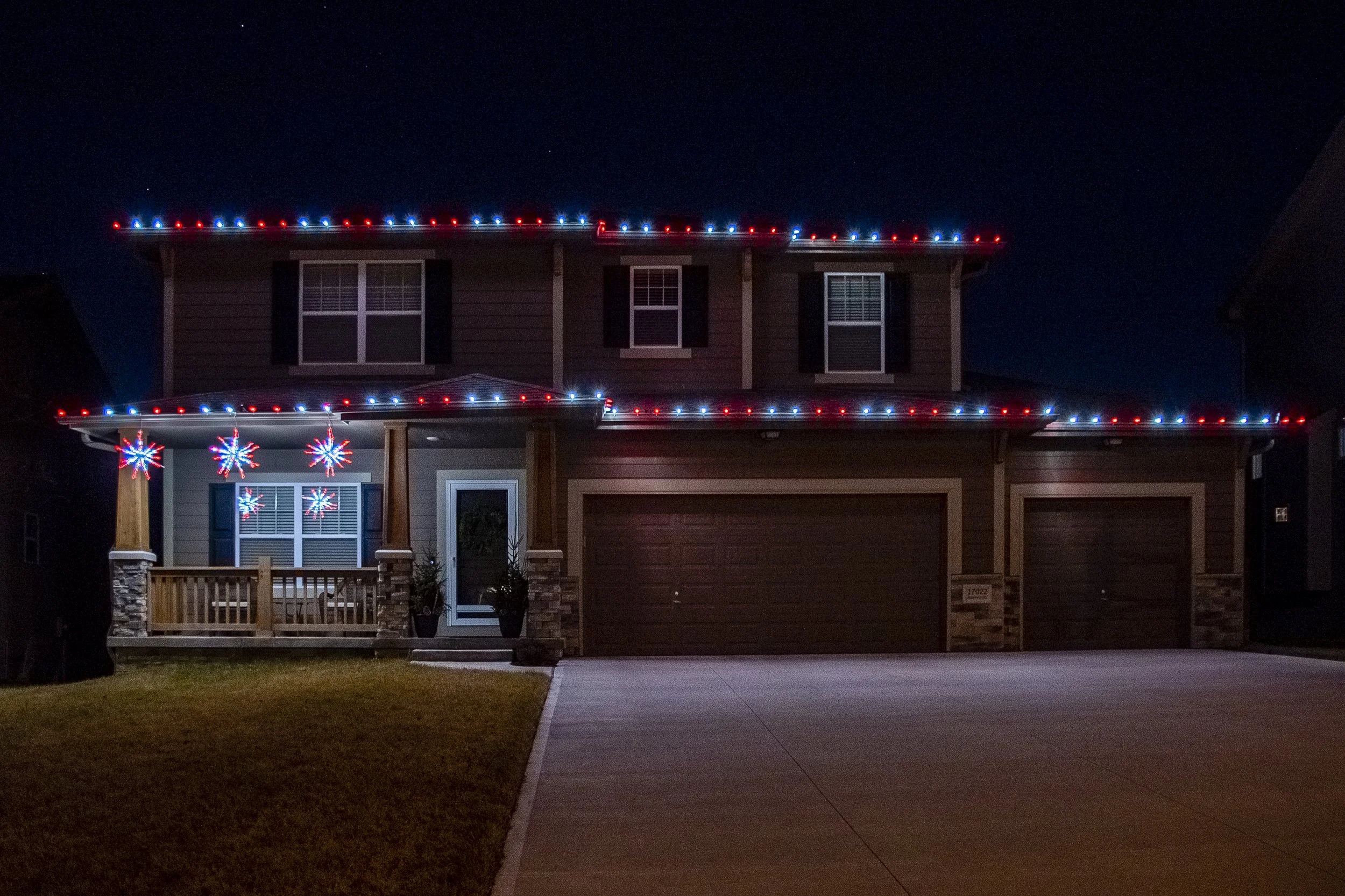 A two-story house decorated with red, white, and blue holiday lights along the roofline, porch, and windows at night.