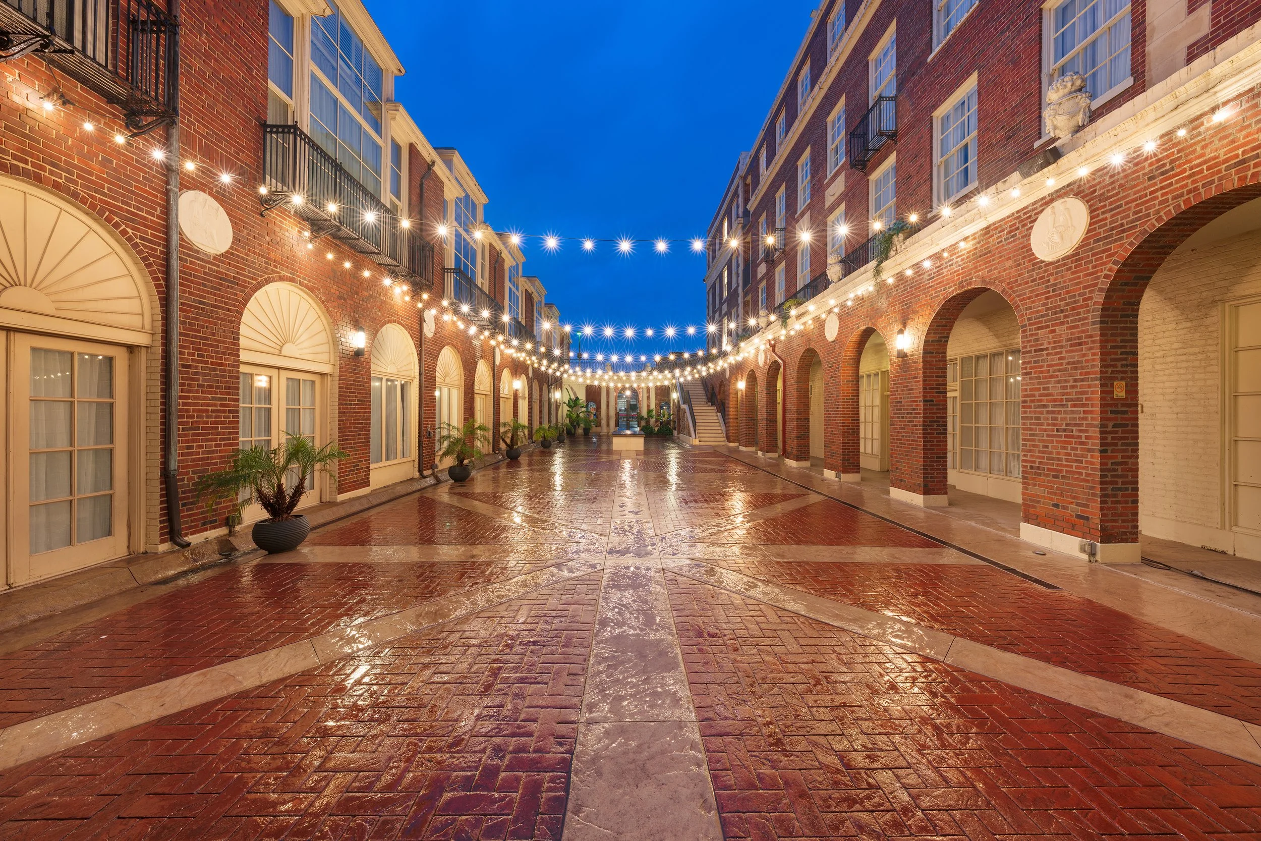 Outdoor courtyard with string lights hanging above, red brick buildings on either side, wet red brick and stone walkway, potted plants along the sides, evening sky in the background.