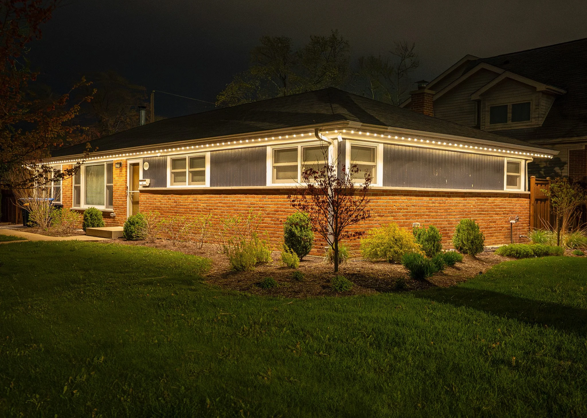 Nighttime photo of a house illuminated by exterior string lights along the roofline, with a well-maintained garden in the foreground and trees in the background.