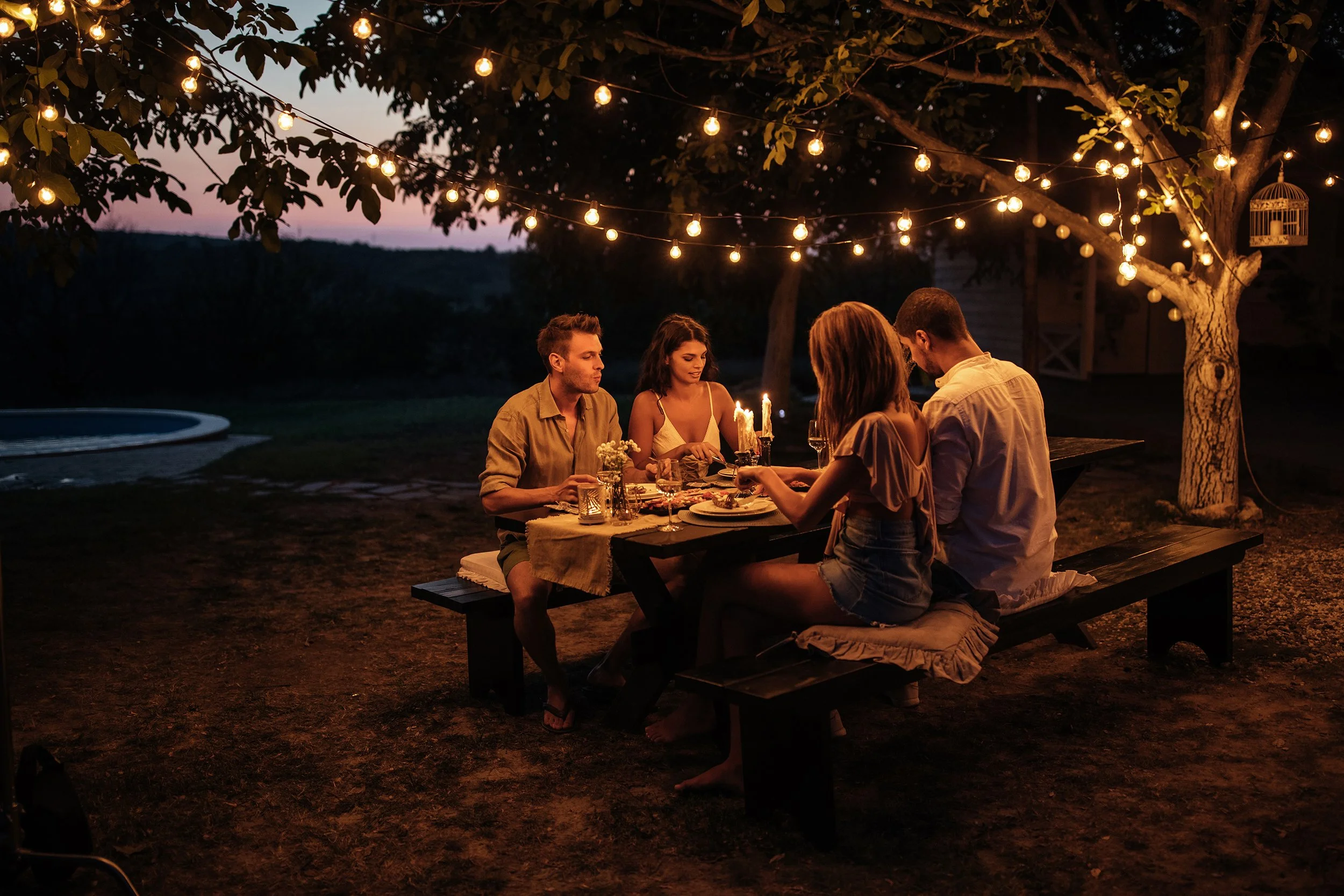 A group of four friends sitting at a picnic table outdoors during evening, under string lights hanging from trees, enjoying dinner together.