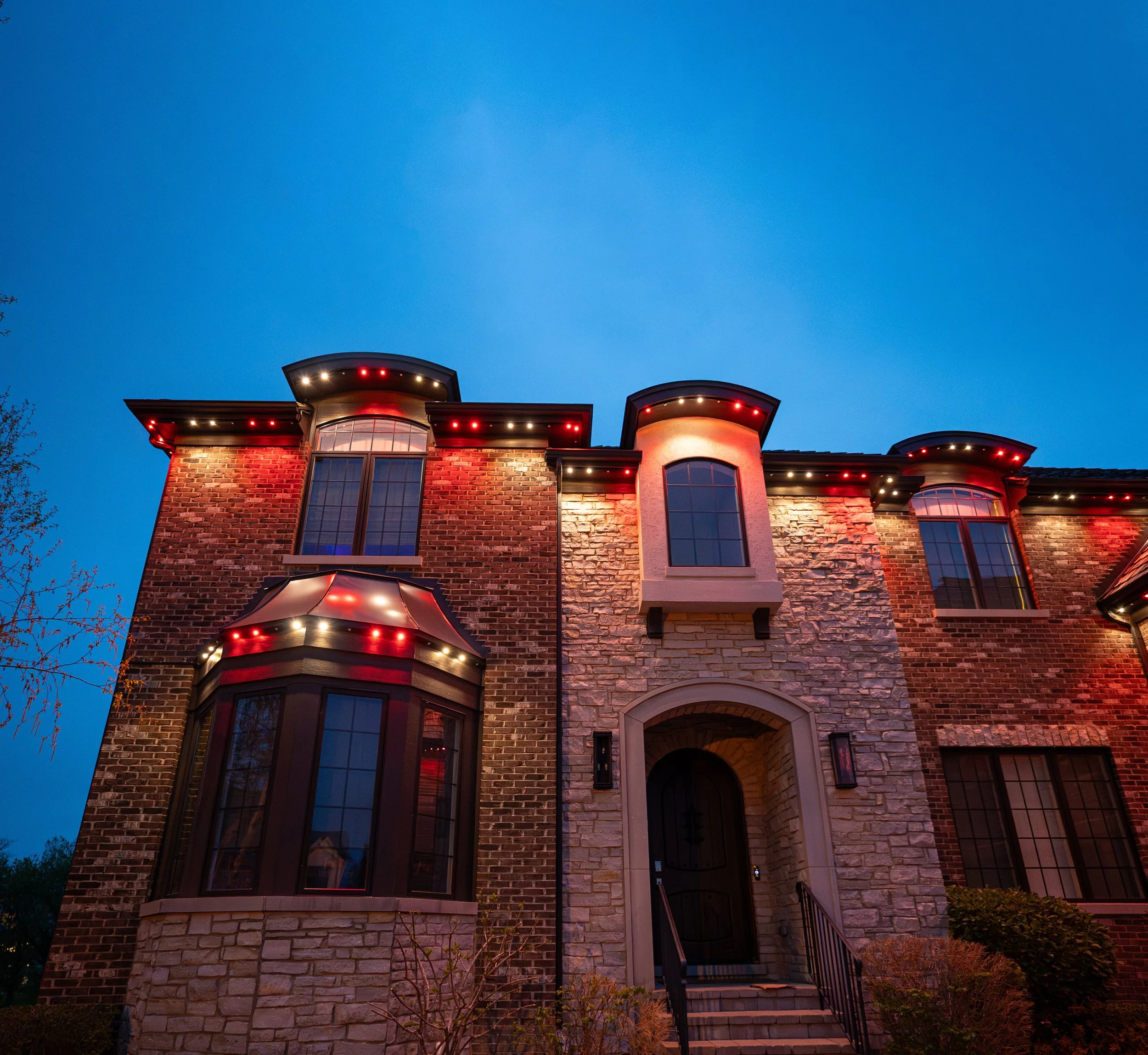 A large brick house at dusk with exterior lighting and window reflections visible.