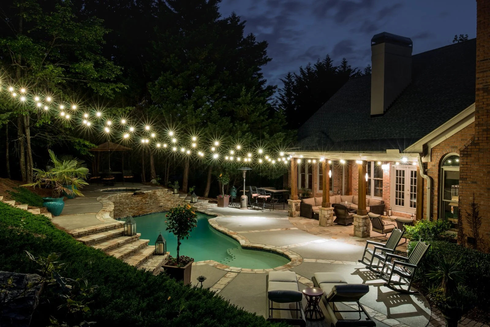Nighttime backyard scene with string lights, pool, patio furniture, and surrounding trees and house.