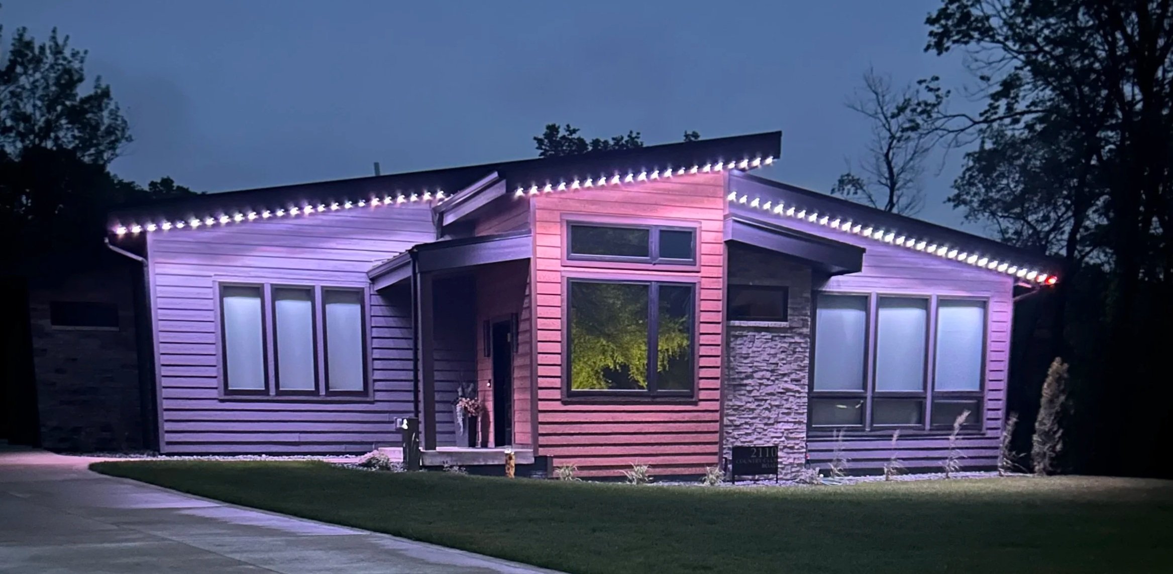 A modern house at dusk illuminated with purple and white multi-colored lights along the roofline, featuring large windows and mixed siding materials, surrounded by trees and a grassy lawn.