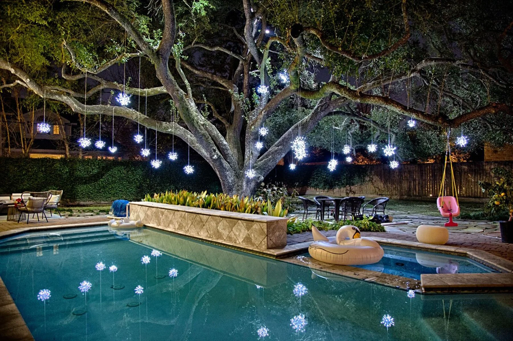 Backyard pool area at night with a large tree decorated with hanging white snowflake-shaped string lights. There are outdoor chairs, a pink swing, pool floaties shaped like swans, and the pool water reflecting the lights.