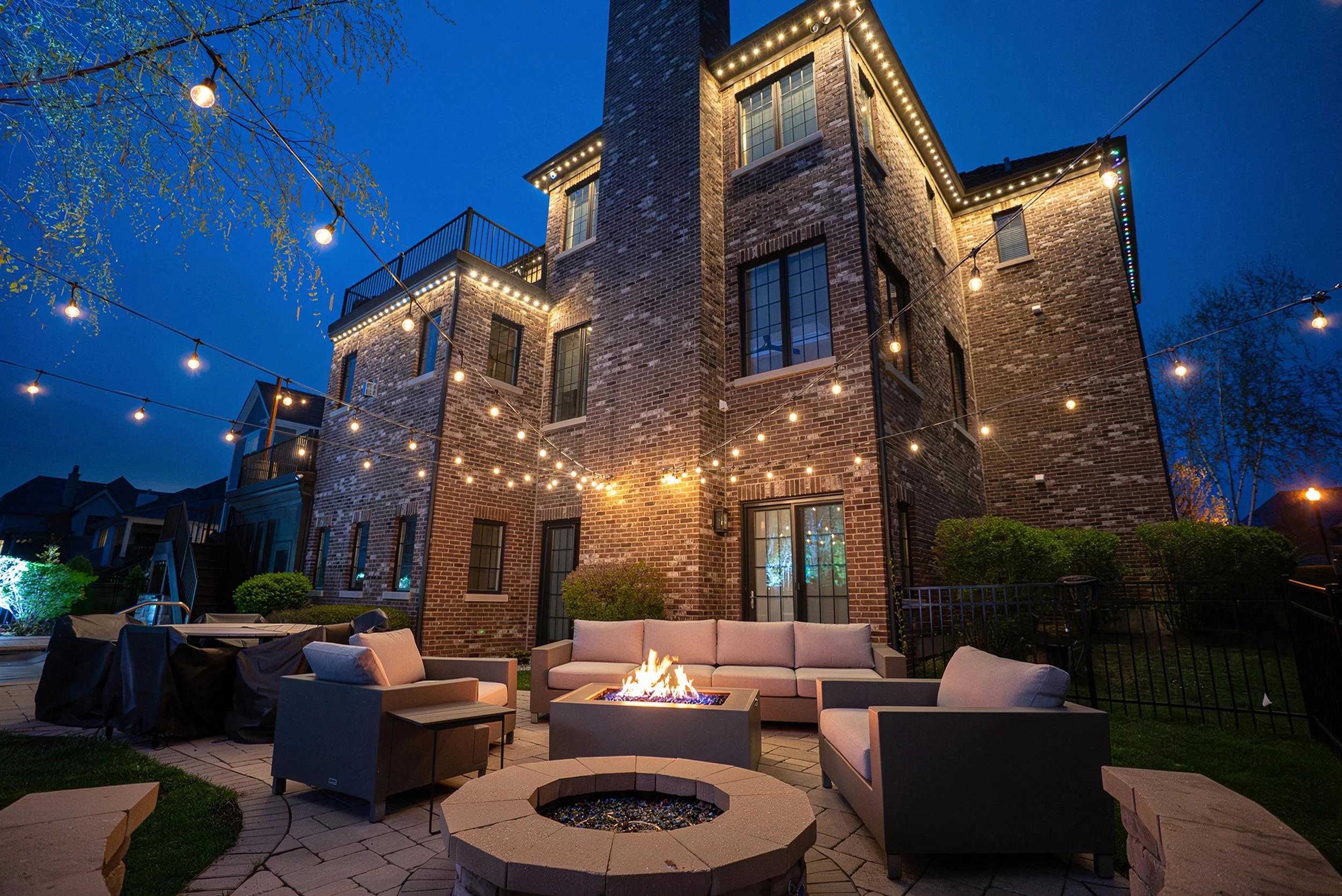 A backyard patio at night featuring a brick house with string lights, outdoor furniture, a fire pit, and a fire in a fire bowl, illuminated under a clear blue sky.