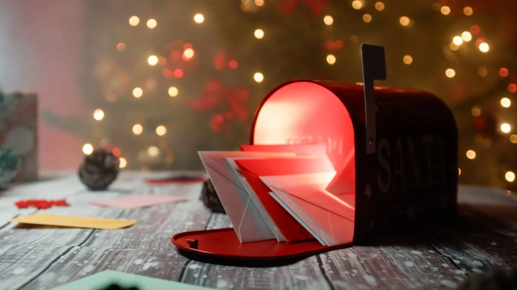 A mailbox with a red light inside, producing a glowing effect, sits on a rustic wooden table with scattered papers and holiday decorations, with a blurred Christmas tree and warm lights in the background.