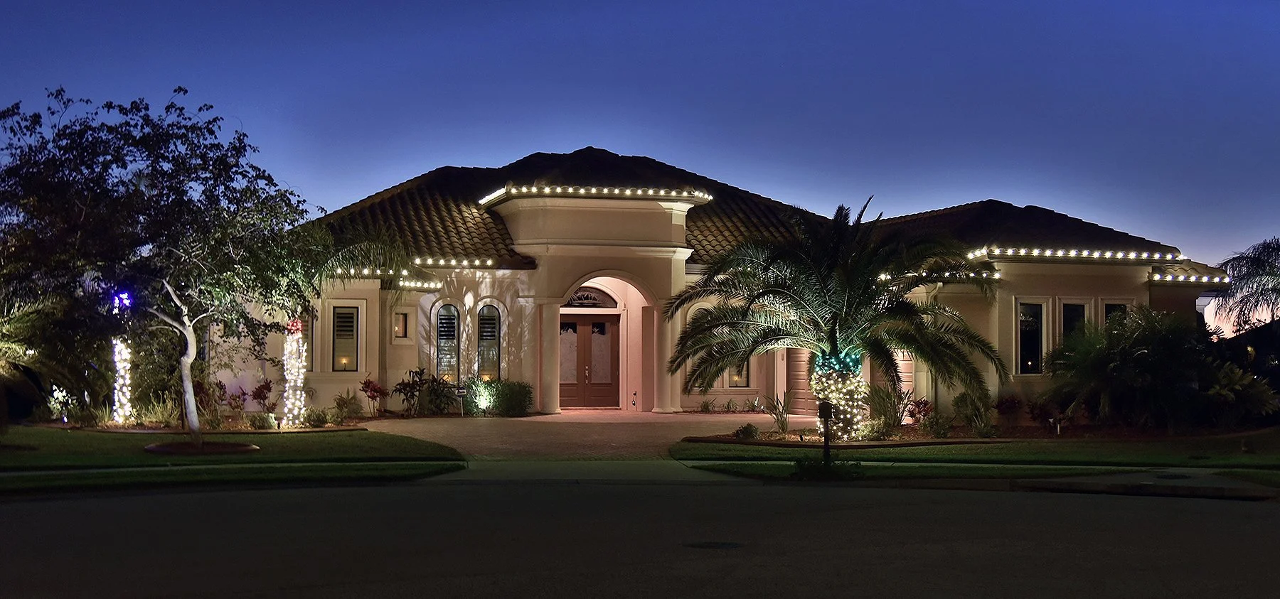 A large, elegant house illuminated with decorative white lights along the roofline and around plants in the yard at dusk or nighttime. The house features a tiled roof, arched windows, a prominent central entrance with double doors, and lush tropical landscaping including palm trees.