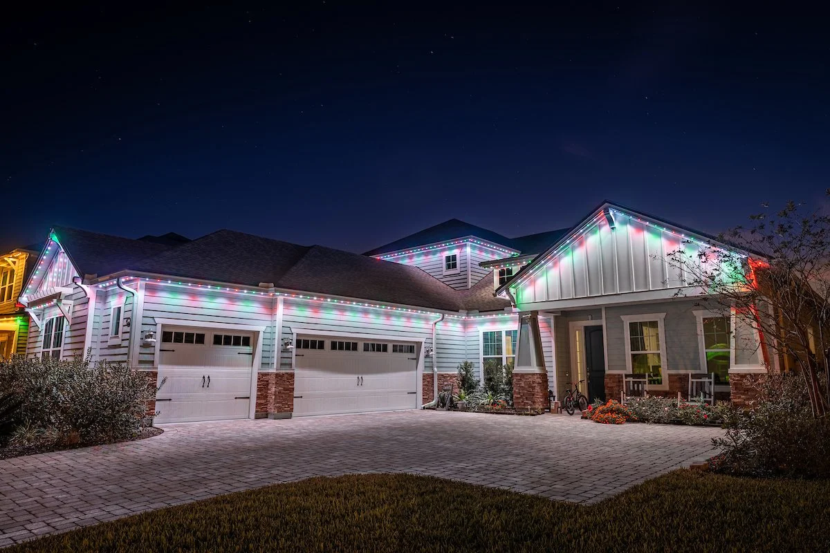 A house decorated with multicolored Christmas lights at night, with a driveway, garage doors, and a small porch area with chairs and bicycles.