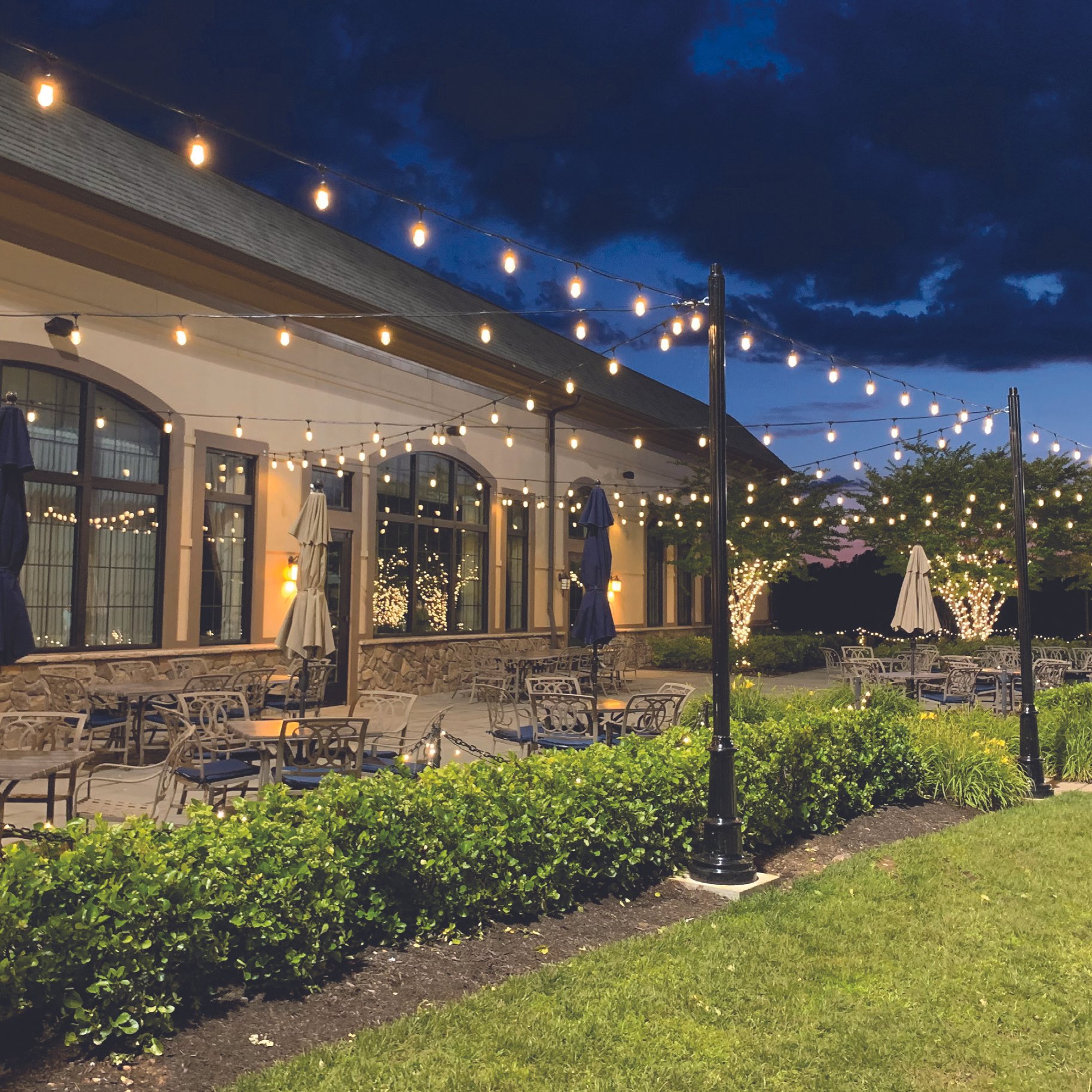 Outdoor patio area decorated with string lights hanging above tables and chairs, with a building featuring large windows and greenery in the background during dusk.