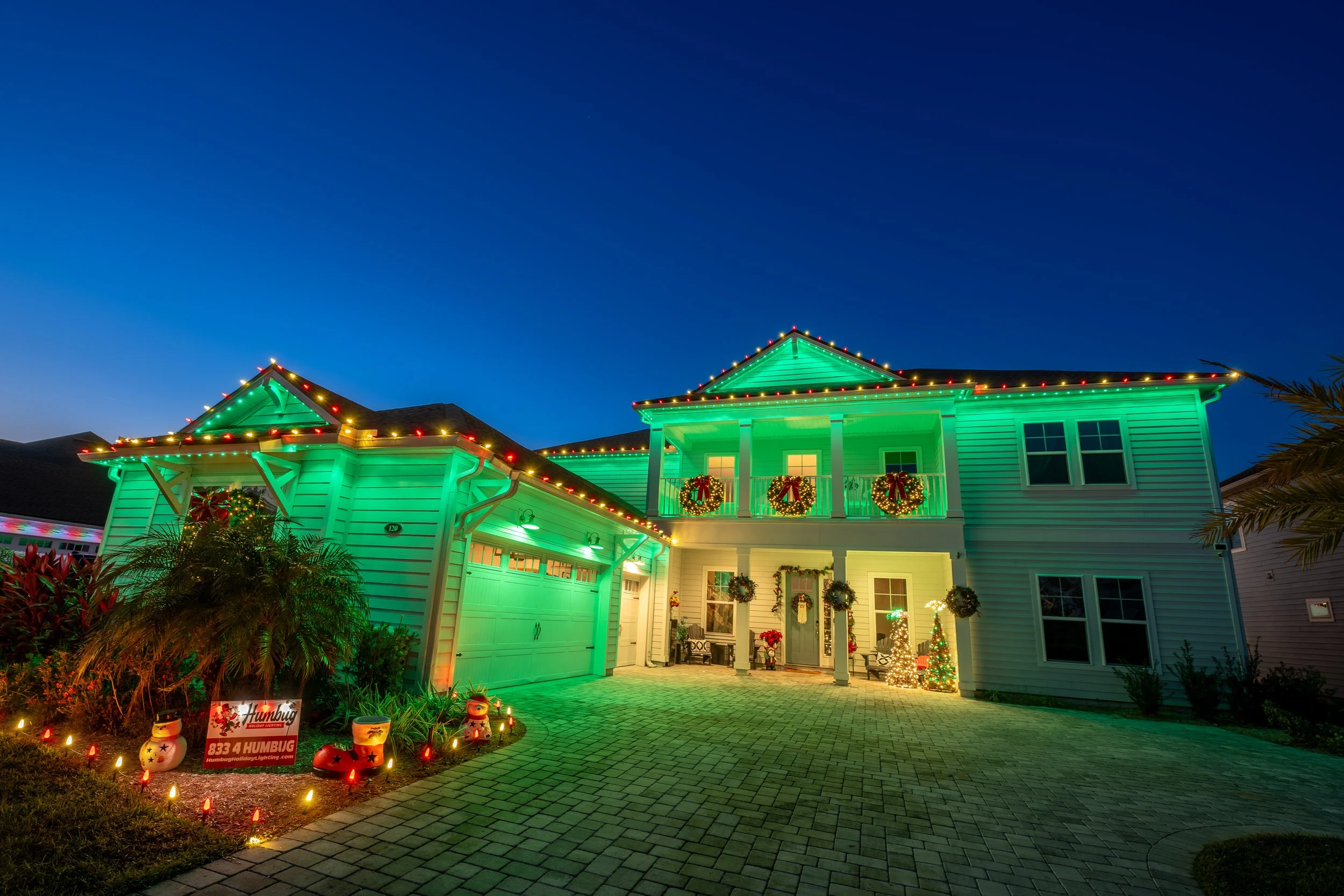 A two-story house decorated with green Christmas lights outline the roof and balcony during evening. The front yard has Christmas wreaths, poinsettias, and a small decorated Christmas tree. There are holiday inflatable decorations and a sign in the garden that reads 'Humbug'. The driveway is paved with bricks, and the house appears festive for Christmas.