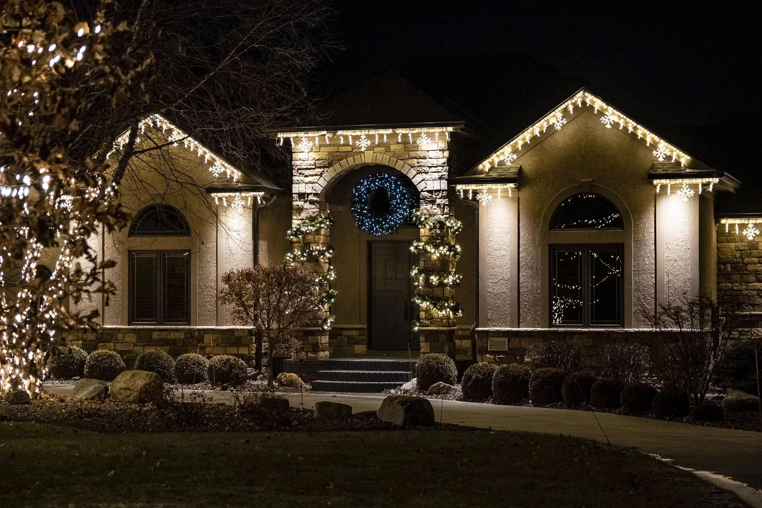 A house decorated with Christmas lights, including icicle lights, snowflake lights, and a festive wreath on the door, illuminated at night.