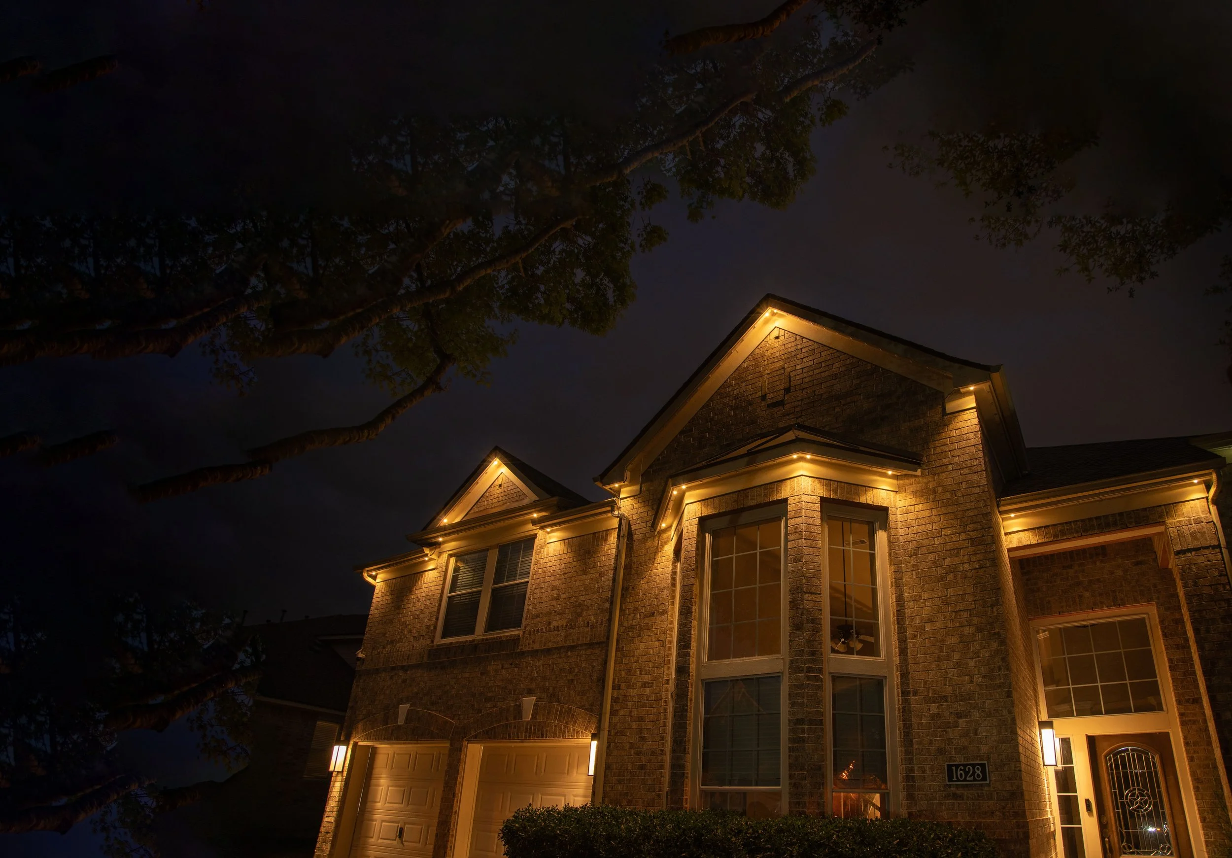 A brick house illuminated with exterior lighting at night, with large windows and a garage. Tall trees are visible in the dark sky.