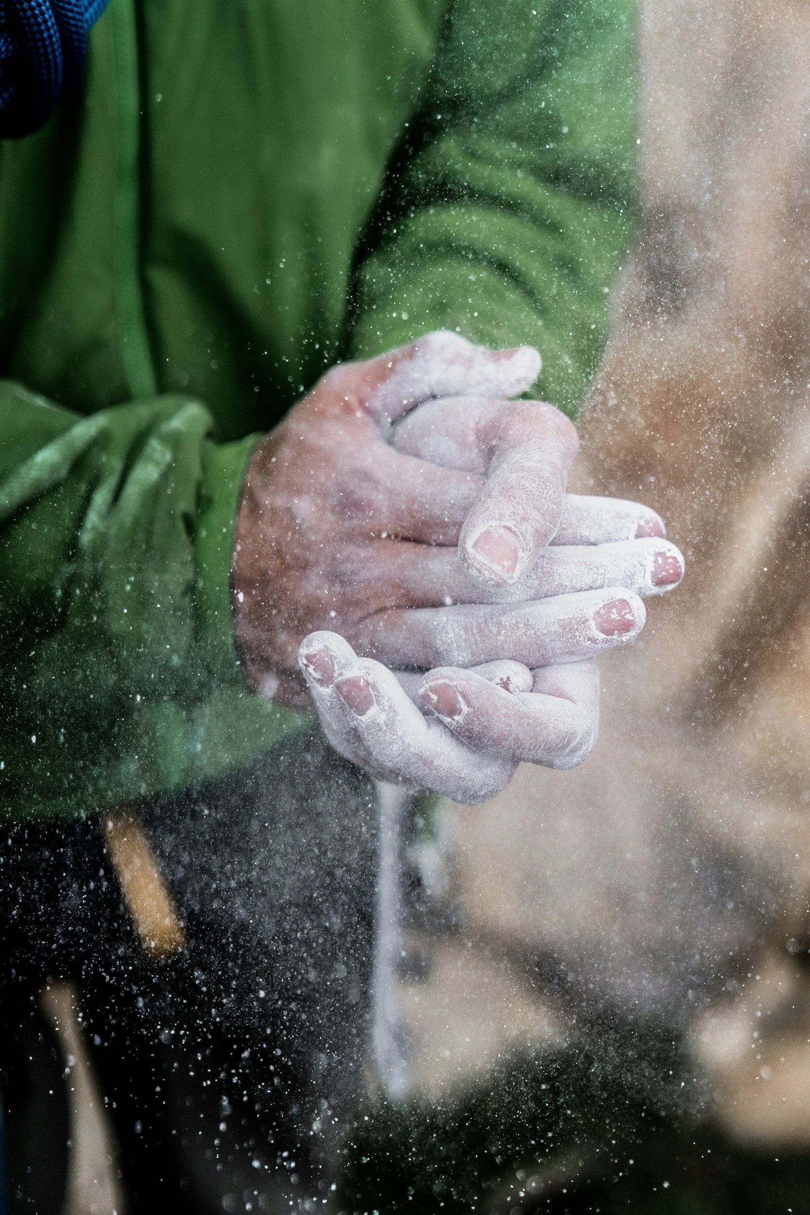 A person wearing a green jacket is dusting their hands with chalk, creating dust particles in the air.