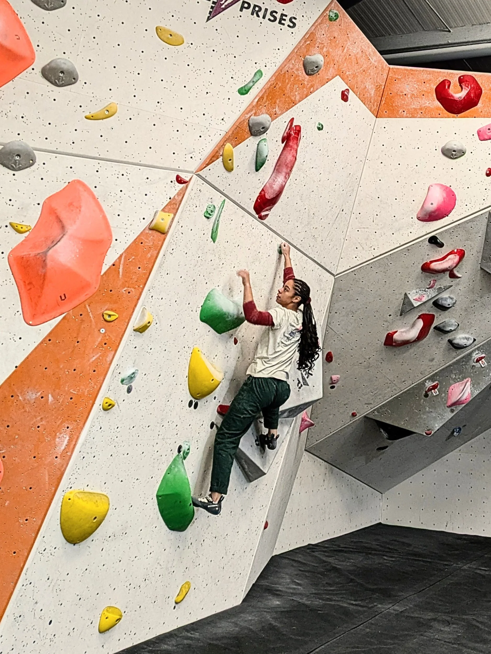 An indoor bouldering climbing gym with various colorful climbing holds on the wall, and a woman with long dark hair climbing.