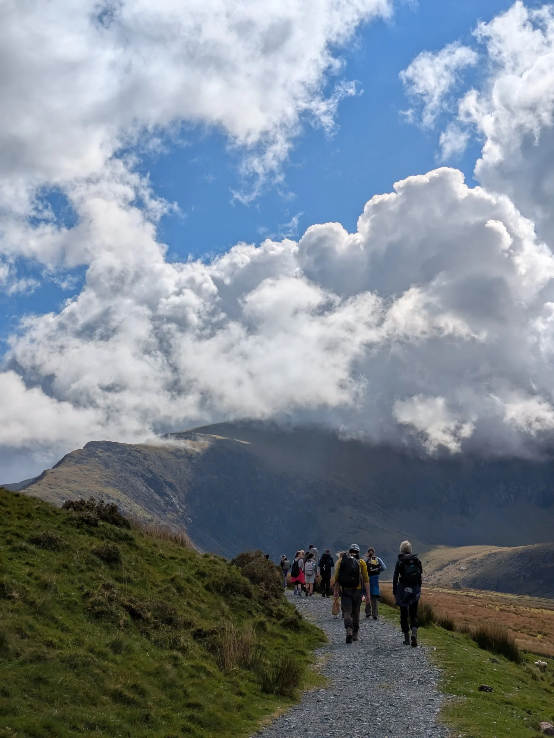 A group of hikers walking on a trail in a mountainous landscape with cloudy blue sky overhead.